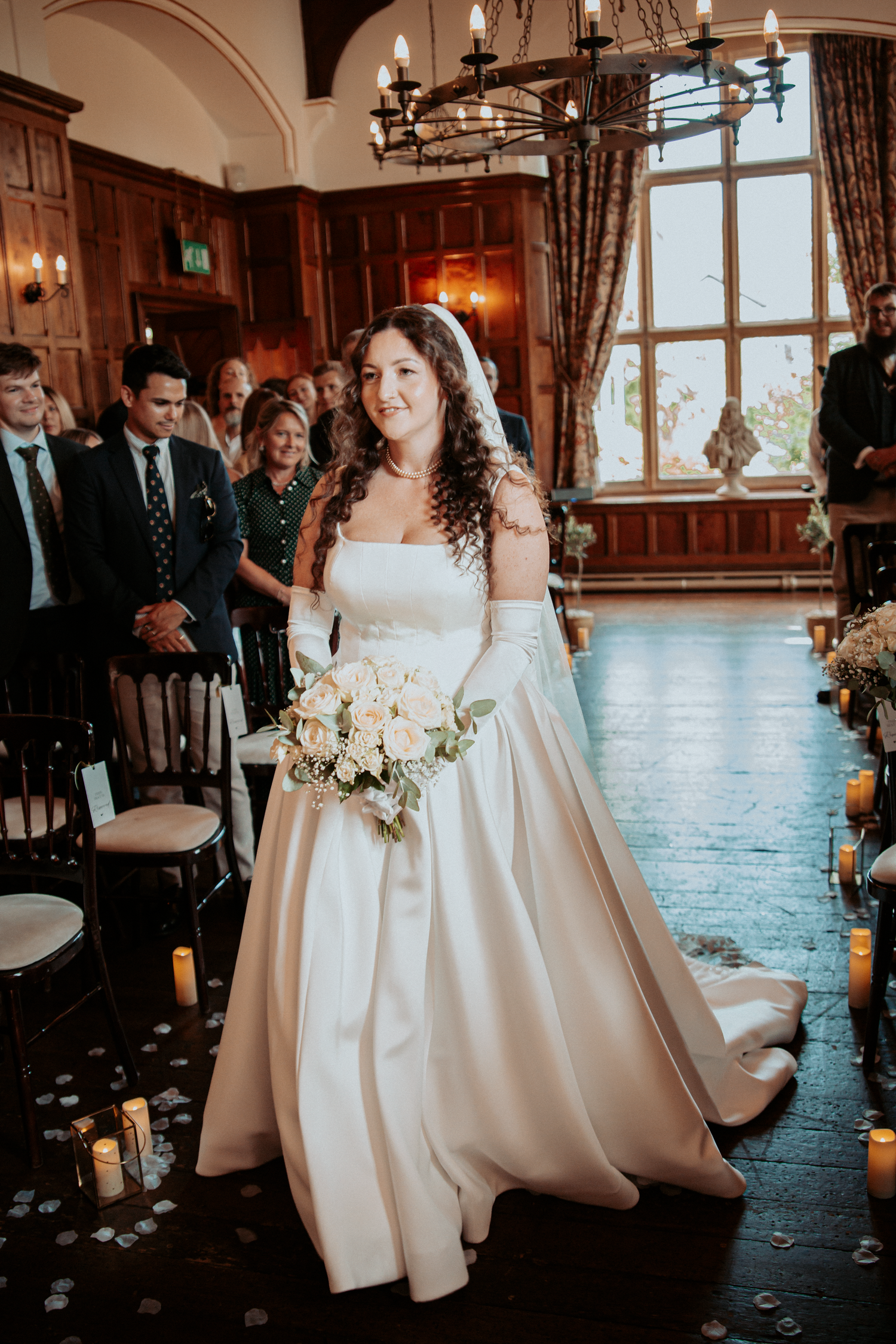 A radiant bride in a white ball gown walks down the aisle holding a bouquet in an elegant wood-paneled venue.