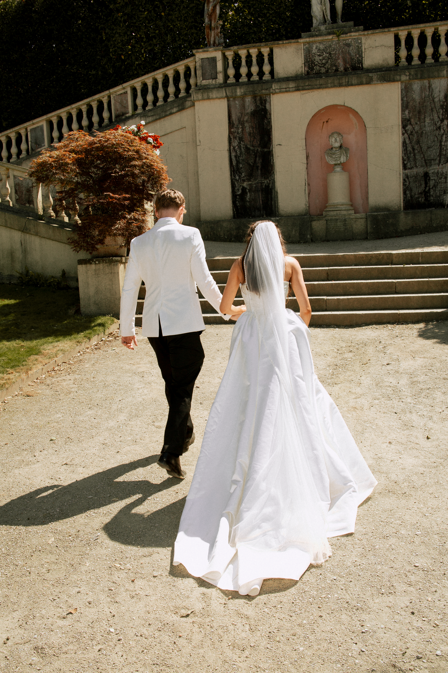 Bride and groom walk hand in hand toward stone staircase in sunlit garden wedding venue.