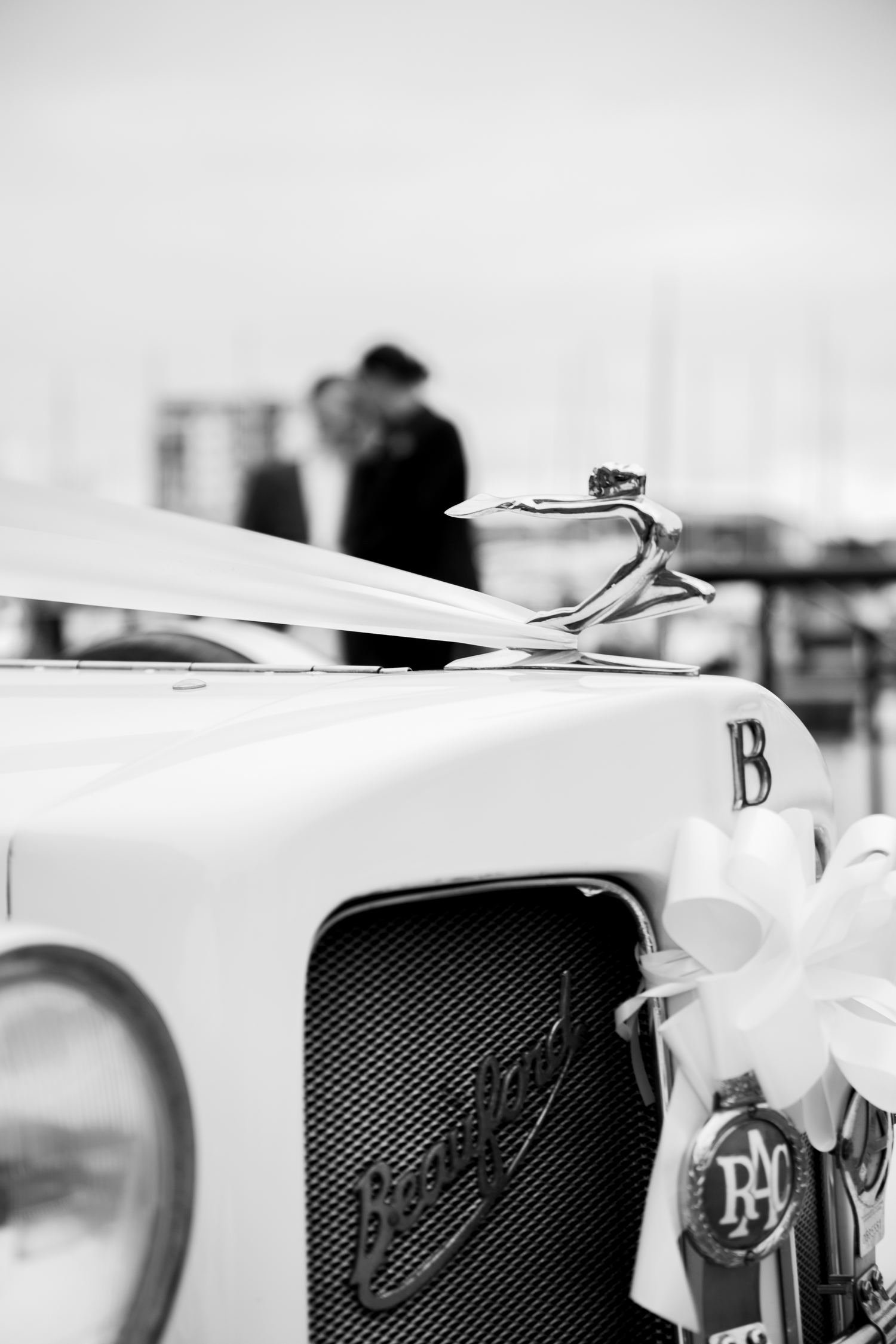 Black and white close-up of a vintage Beauford wedding car hood ornament with couple kissing in background.