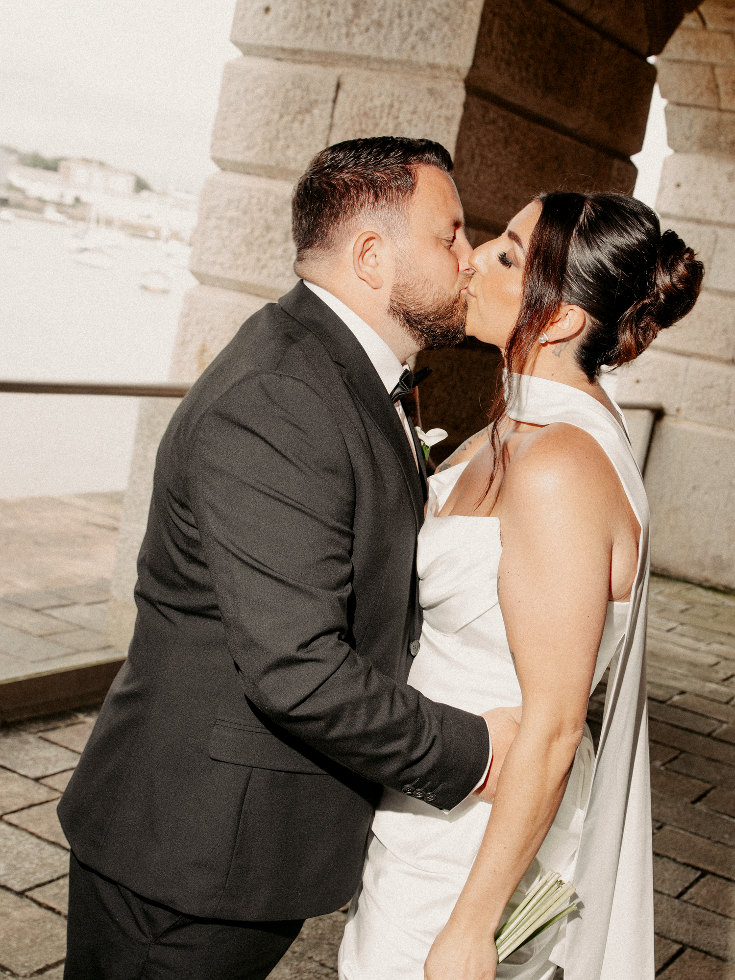 Bride and groom kissing near stone arch columns, bride in white halter gown, groom in dark suit.