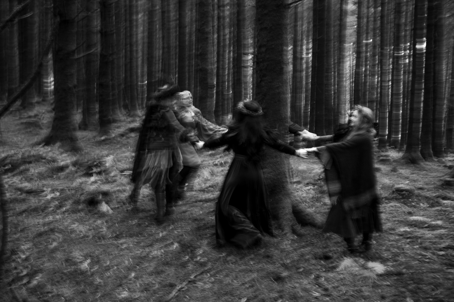 Black and white blurred motion shot of people in dark cloaks dancing in a forest.