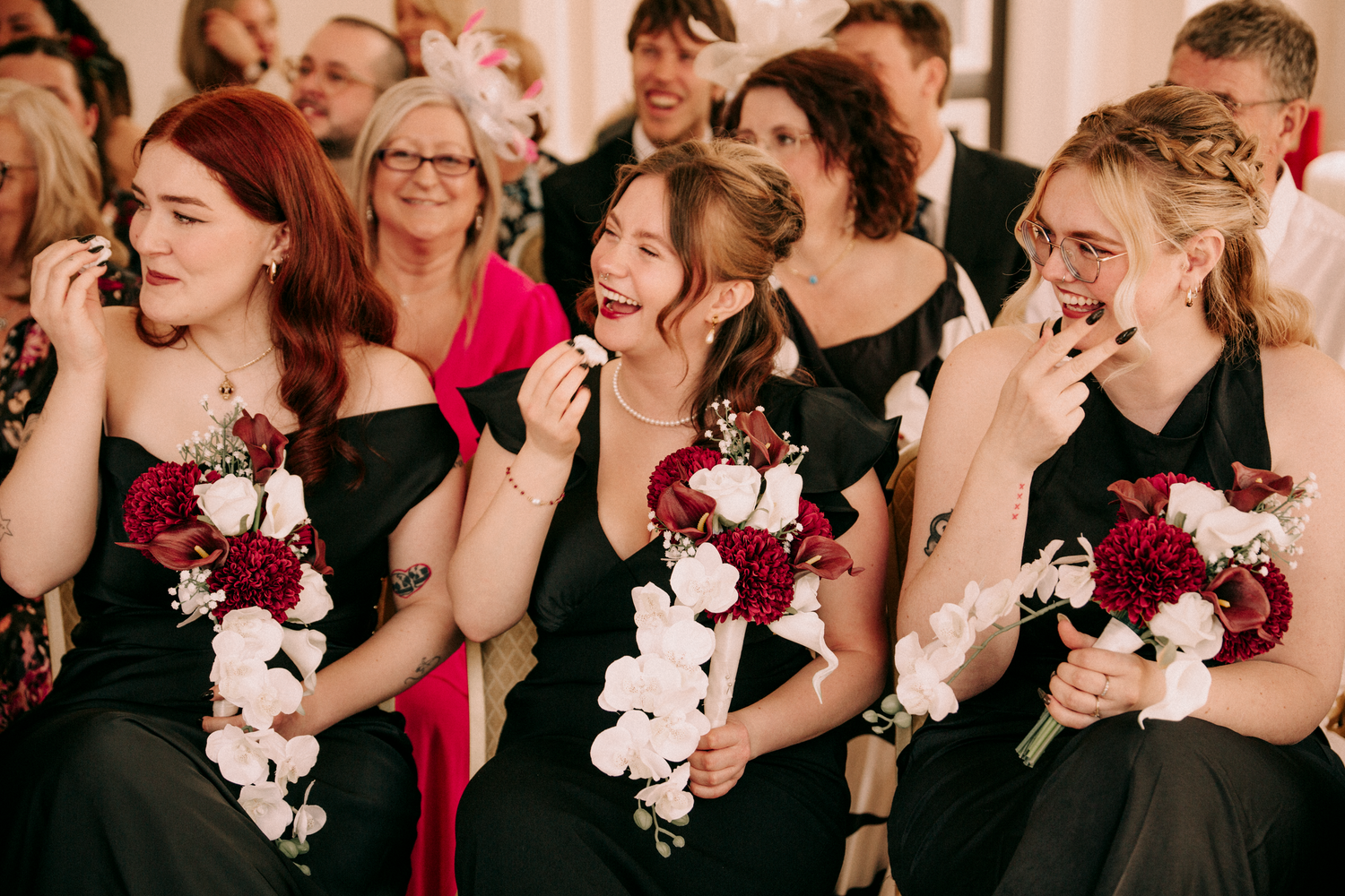 Bridesmaids in black dresses holding red and white bouquets laughing emotionally during wedding ceremony.
