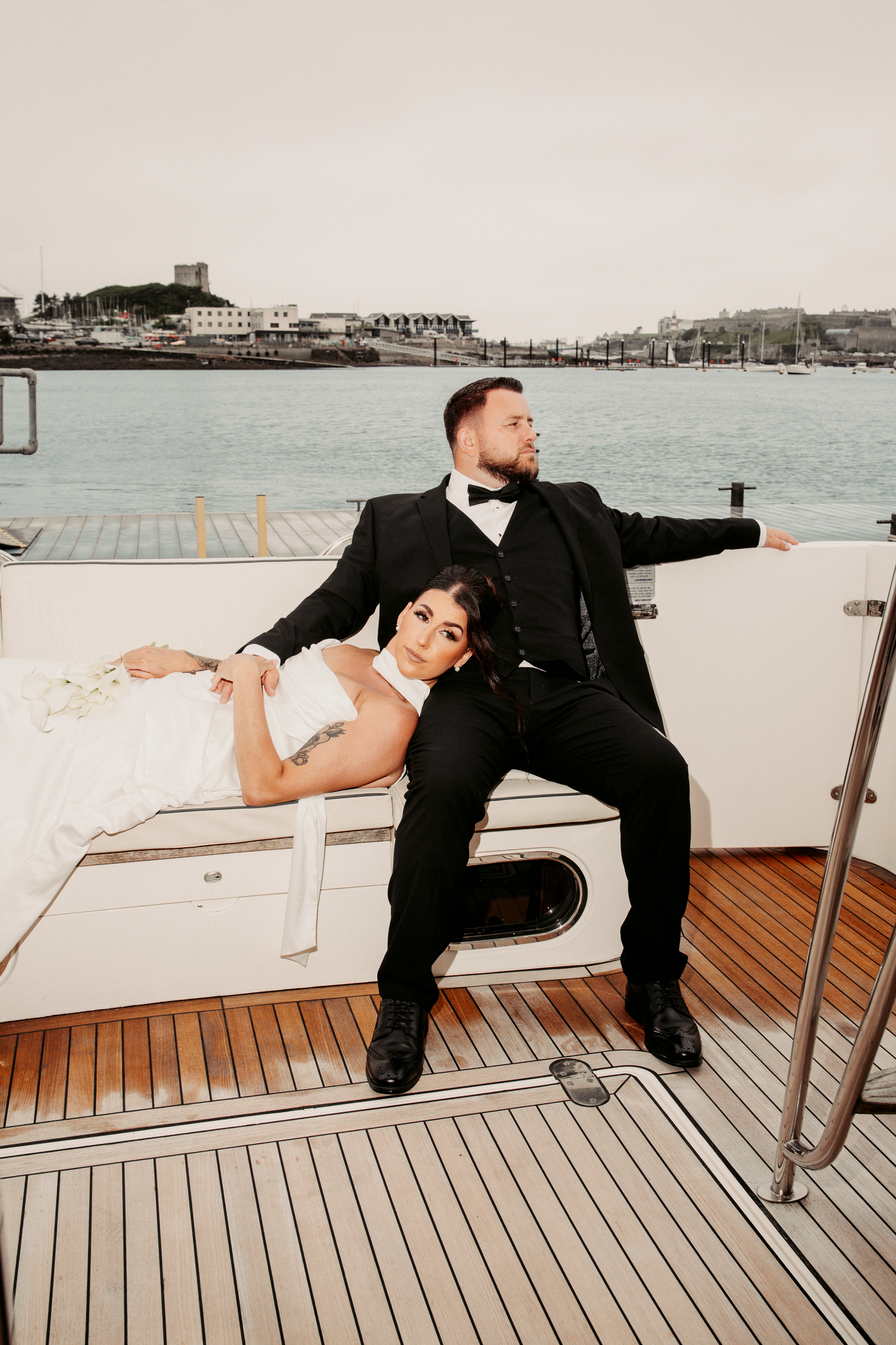 Bride reclining against groom seated on yacht deck, both in formal wedding attire by the harbor.