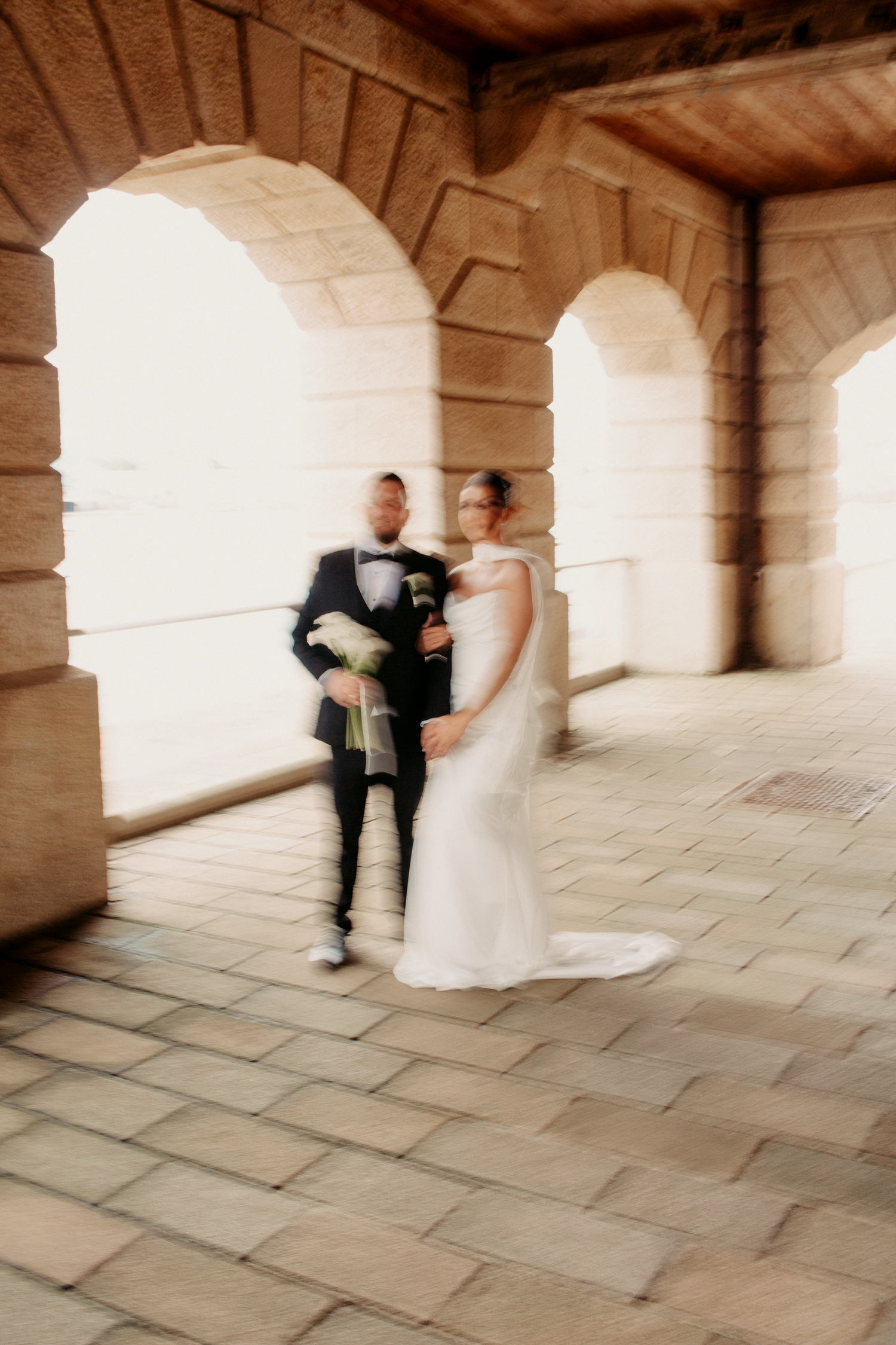 Bride and groom pose under stone archways in a softly blurred artistic wedding portrait outdoors.