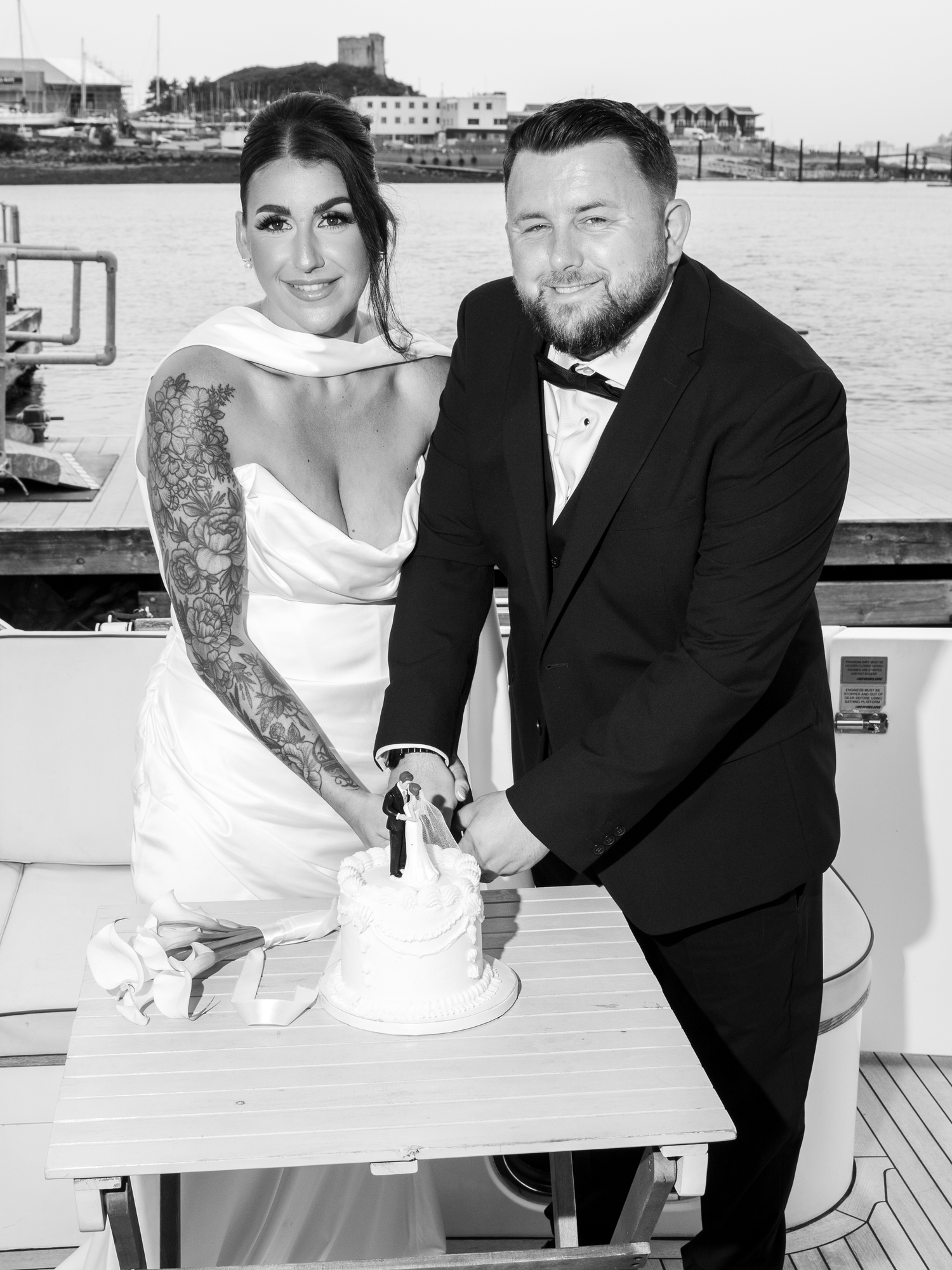 Black and white photo of bride and groom cutting wedding cake on a yacht with waterfront backdrop.