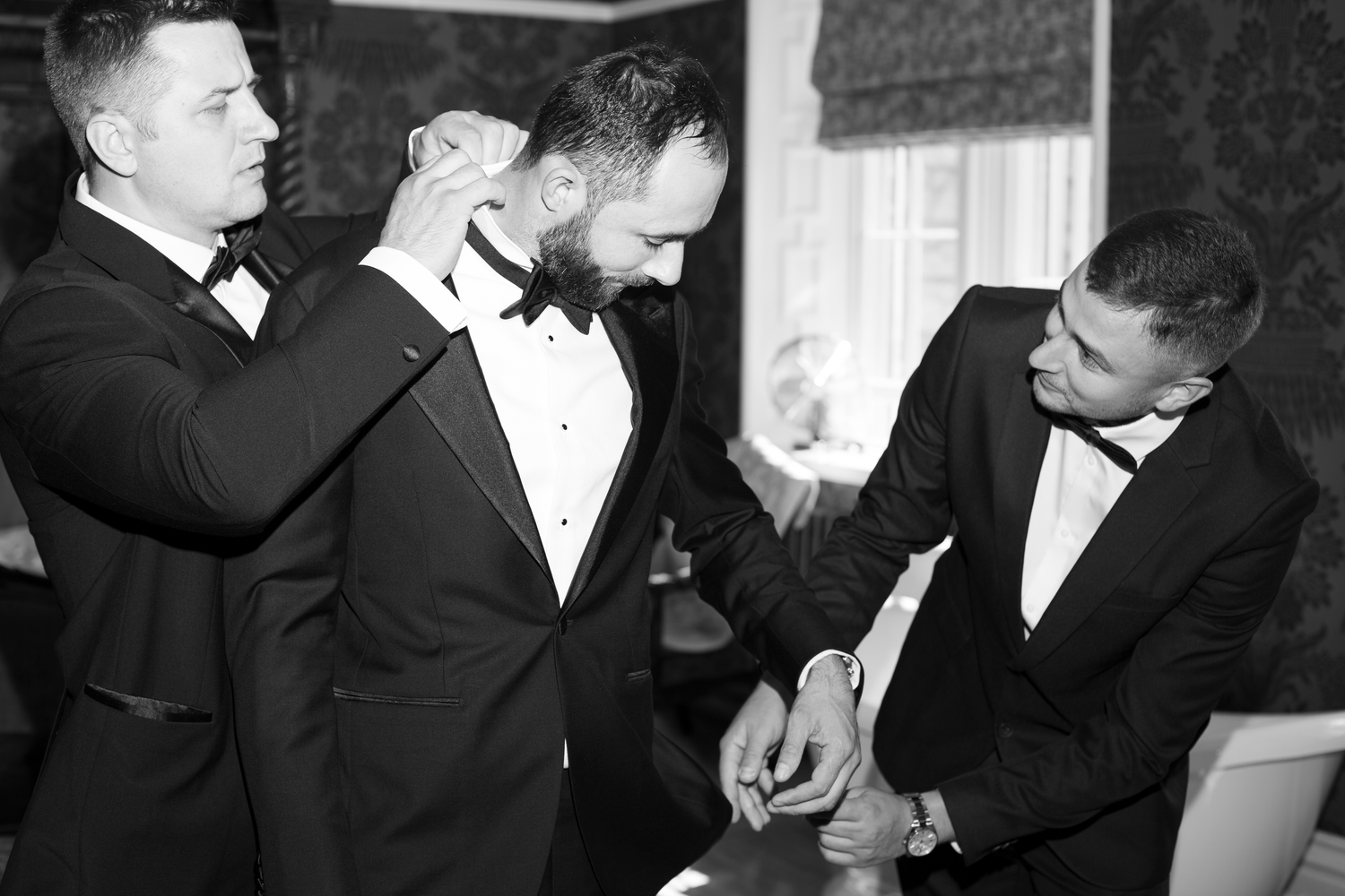 Black and white photo of groomsmen helping groom adjust his tuxedo bow tie before wedding ceremony.