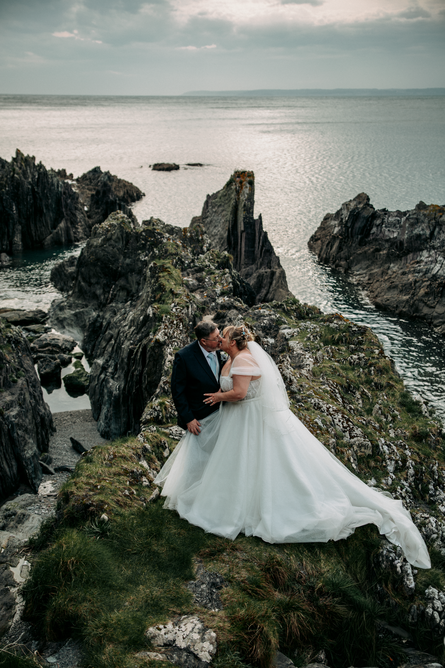 Bride and groom share a kiss on dramatic coastal cliffs overlooking the ocean at sunset.