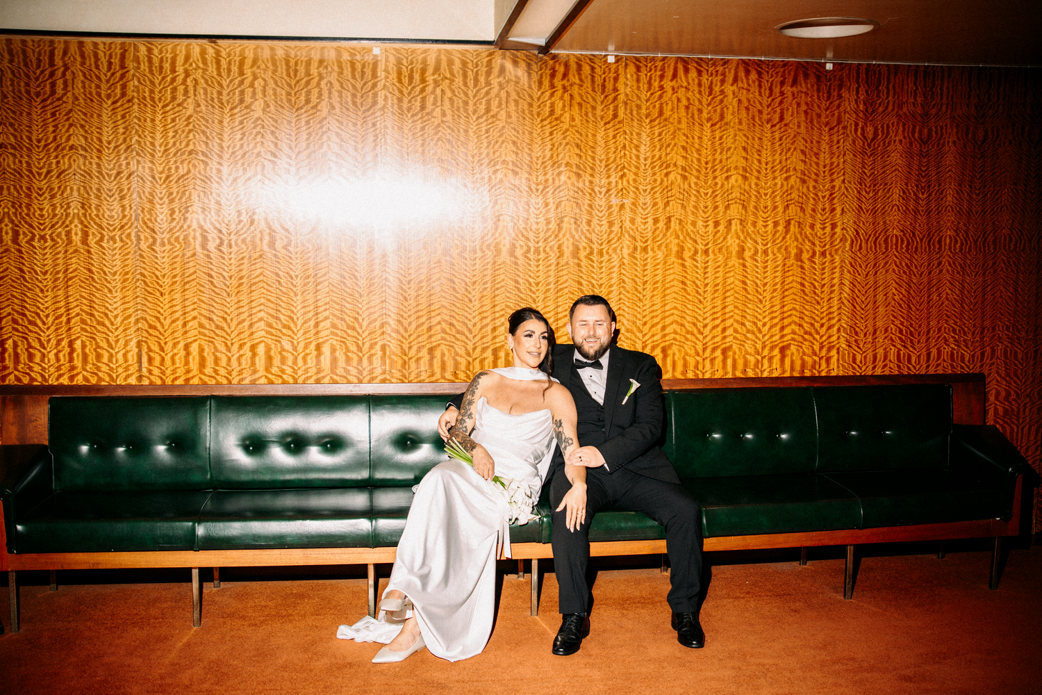 Bride and groom sit together on a green leather sofa in a mid-century modern styled wedding venue.