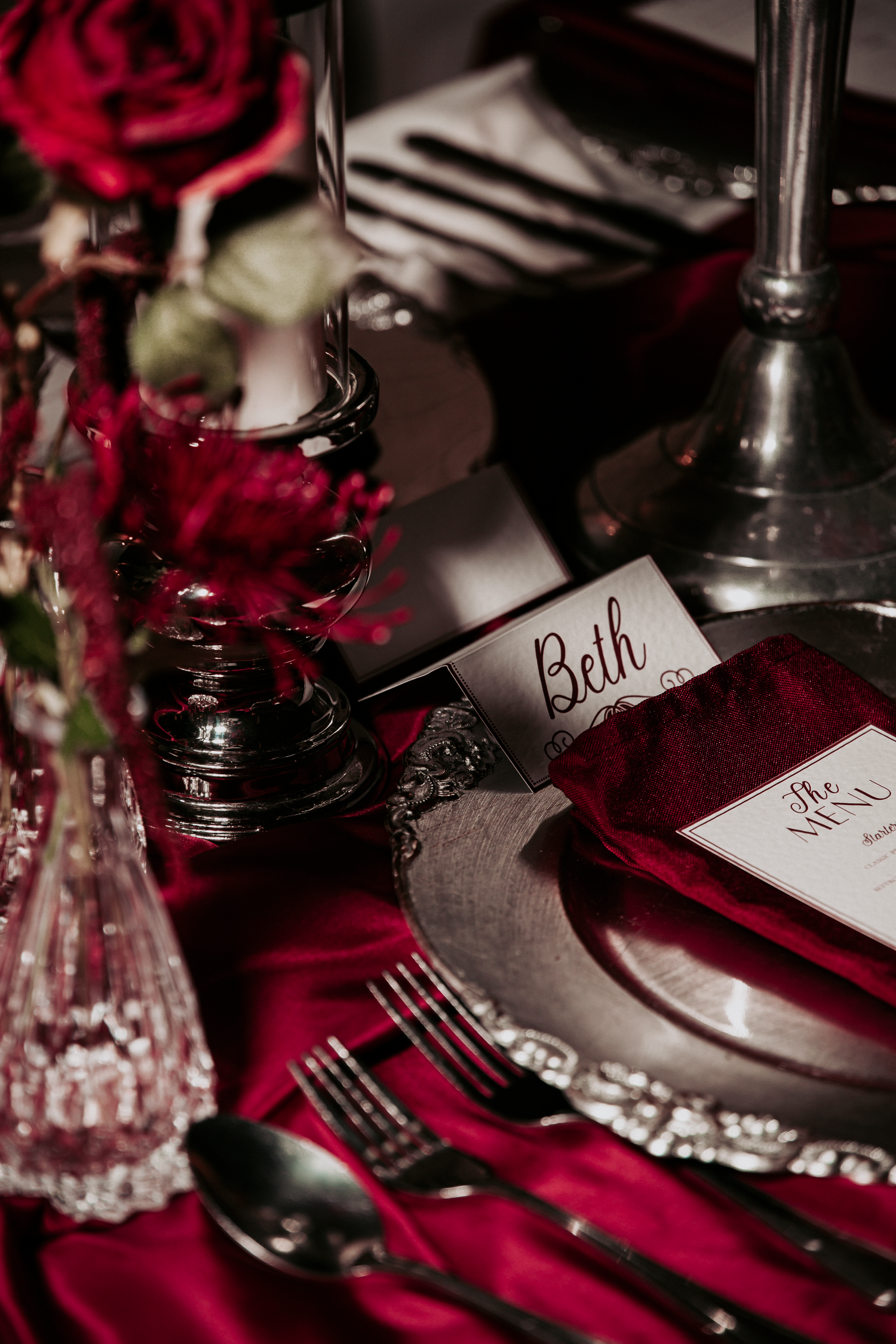 Elegant dark red wedding table setting with roses, silver candelabras, velvet napkins, and a personalized place card reading Beth.