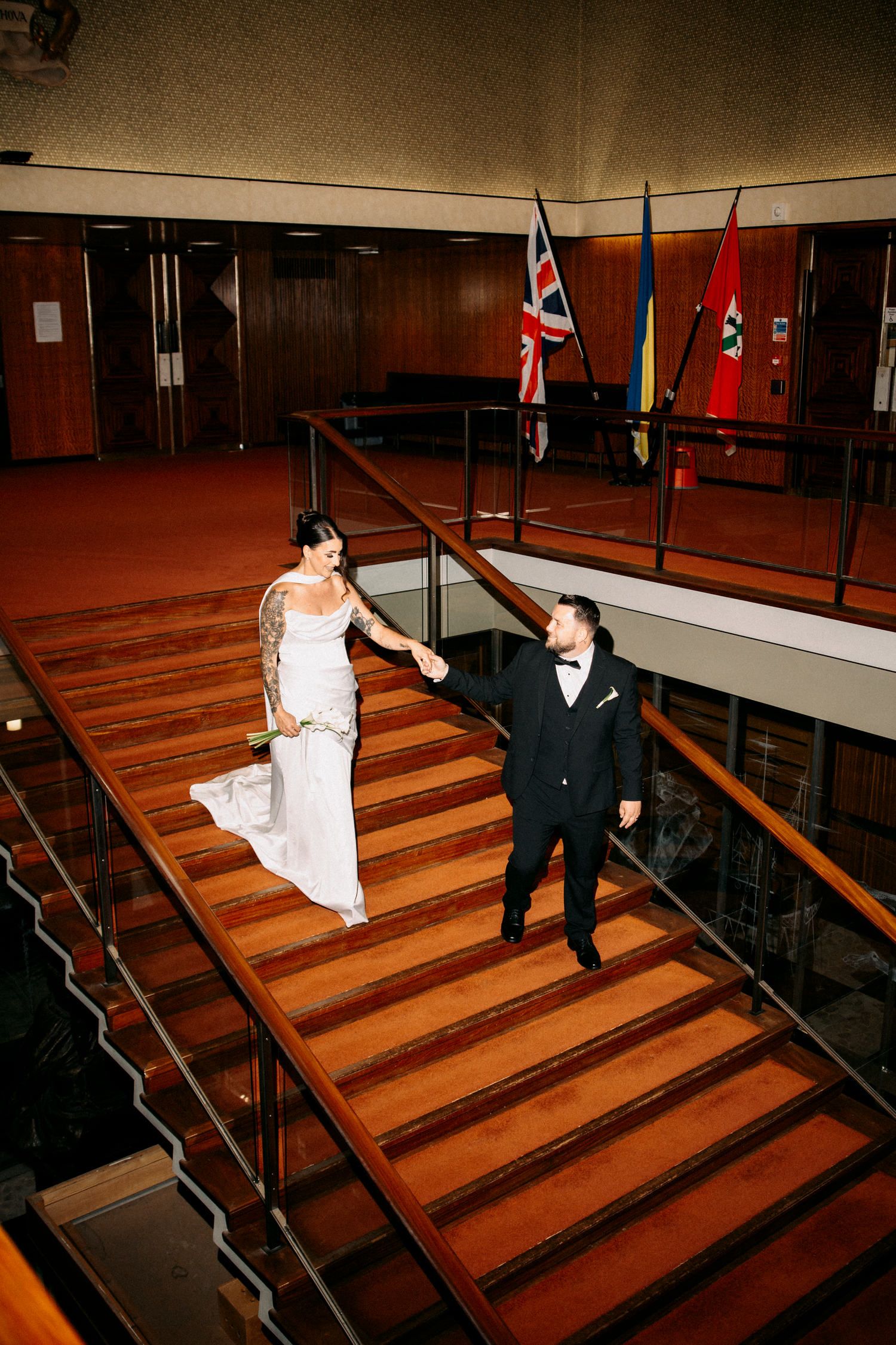 Bride and groom holding hands while descending wooden stairs in an elegant venue with flags.