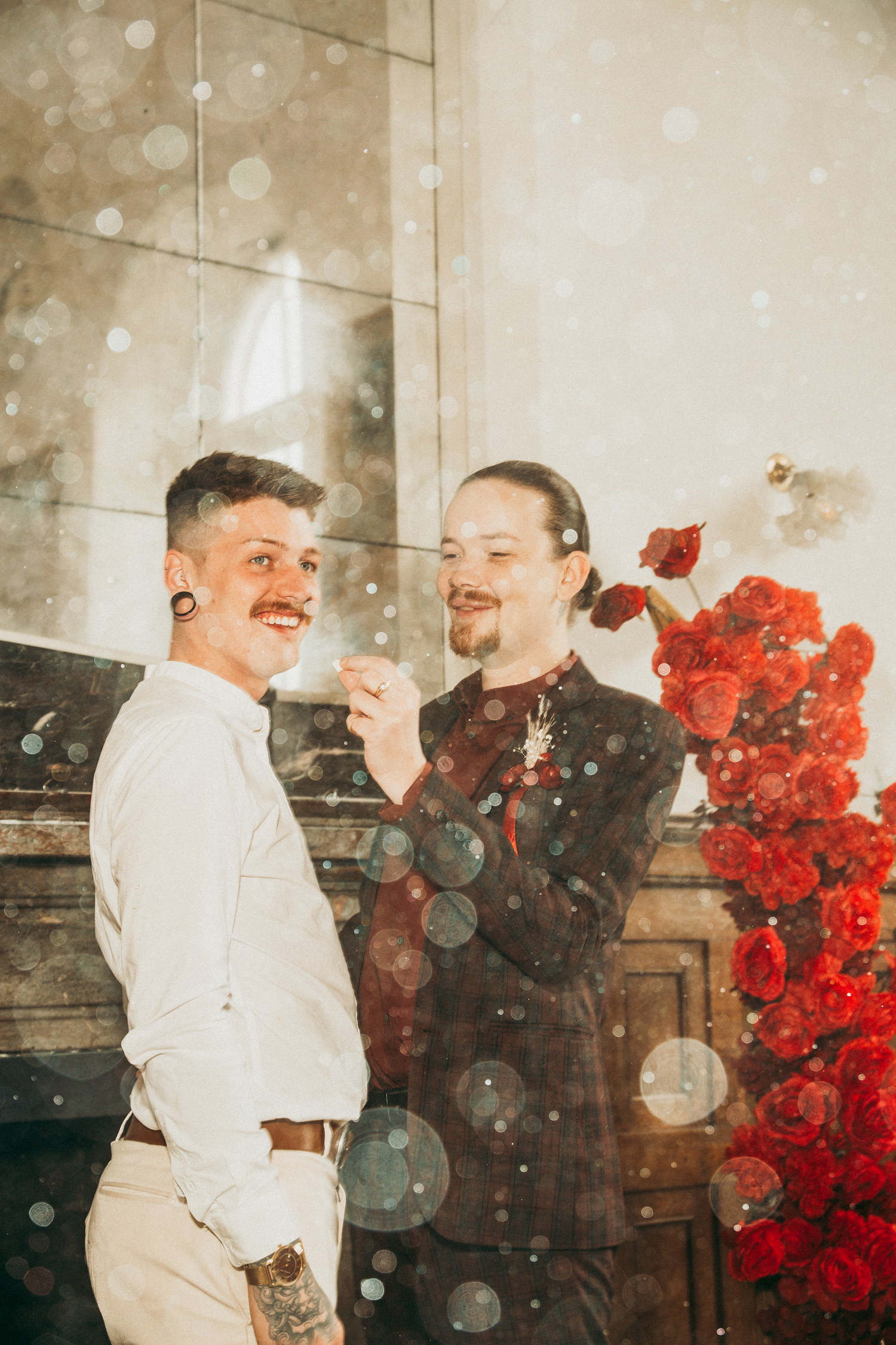 Two grooms sharing joyful moment with sparkler near red roses, dreamy bokeh double exposure effect.