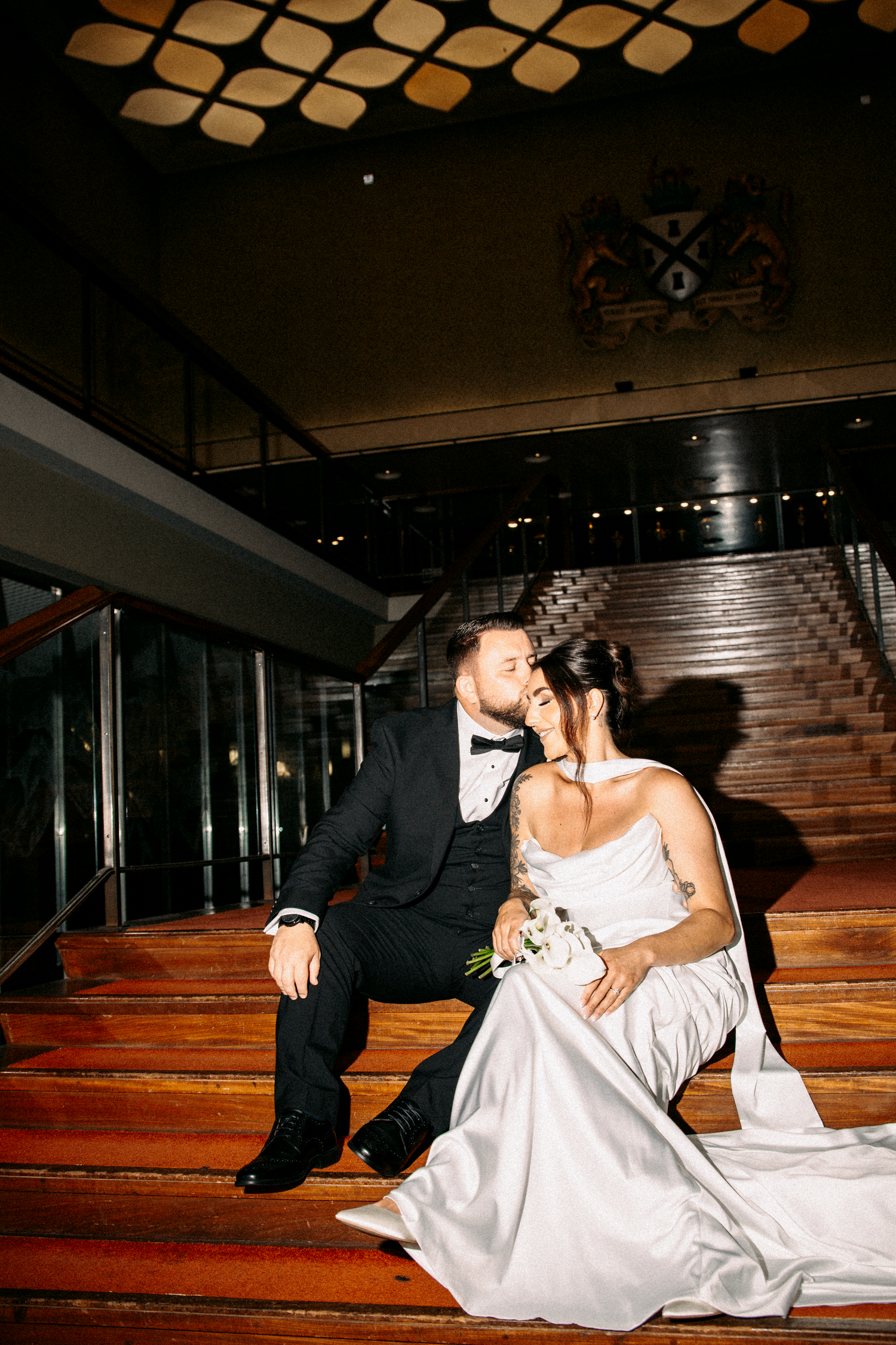 Wedding couple sitting intimately on carpeted stairs, groom in black tuxedo leaning toward bride in strapless white gown.
