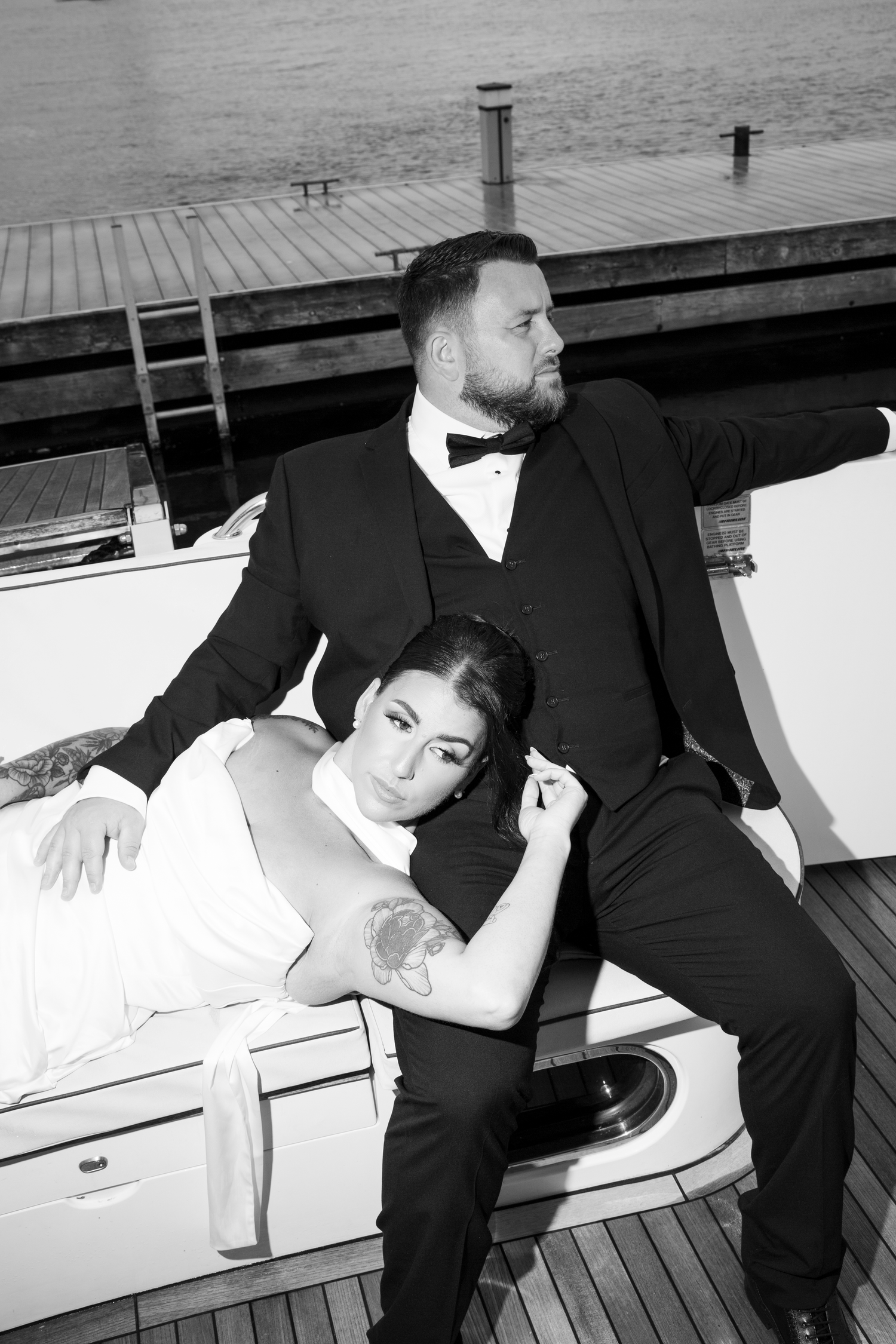 Bride resting on groom's lap on a yacht deck, both in wedding attire, elegant black and white portrait.