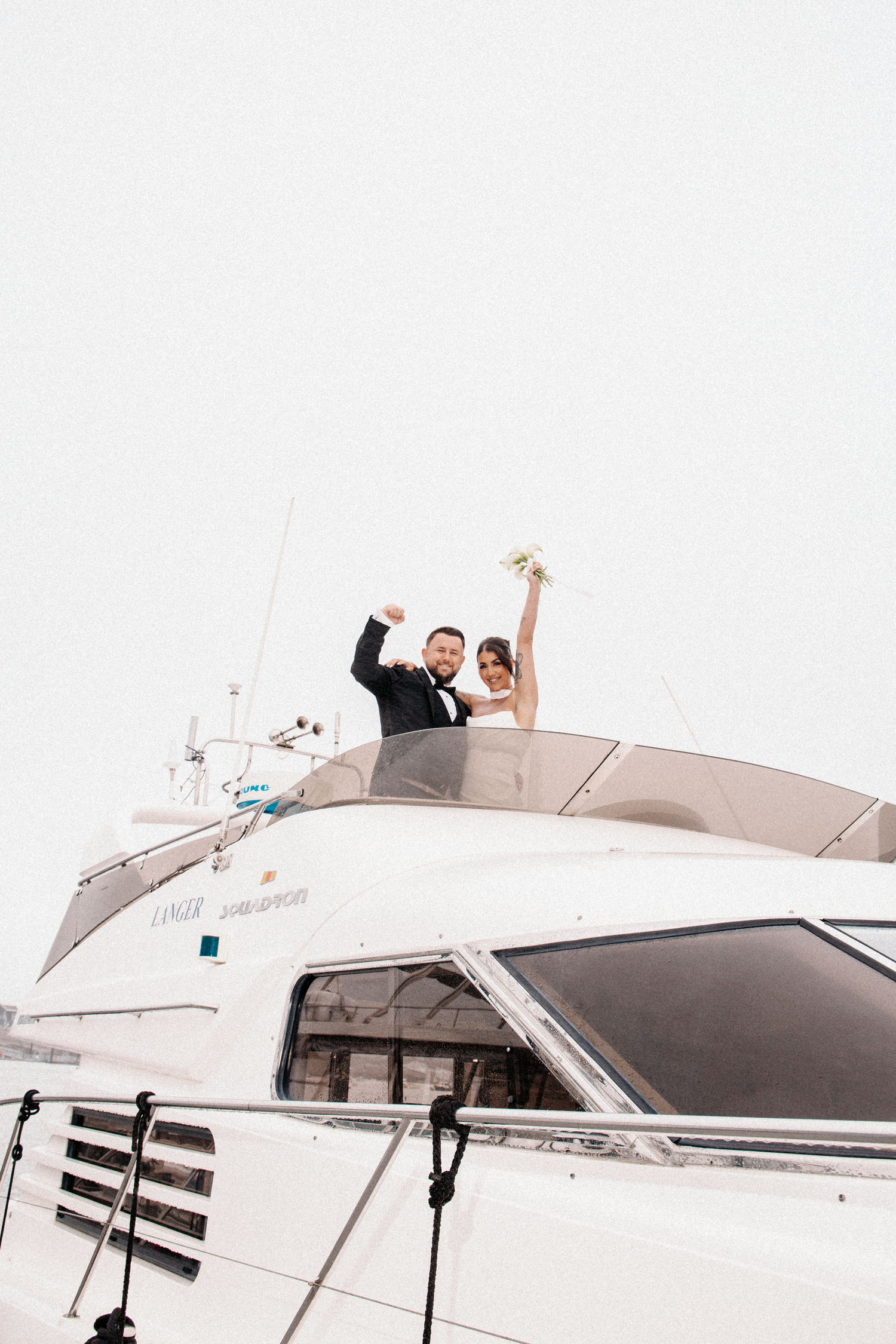Joyful bride and groom celebrating on top of a luxury yacht, raising arms with bouquet in the air.