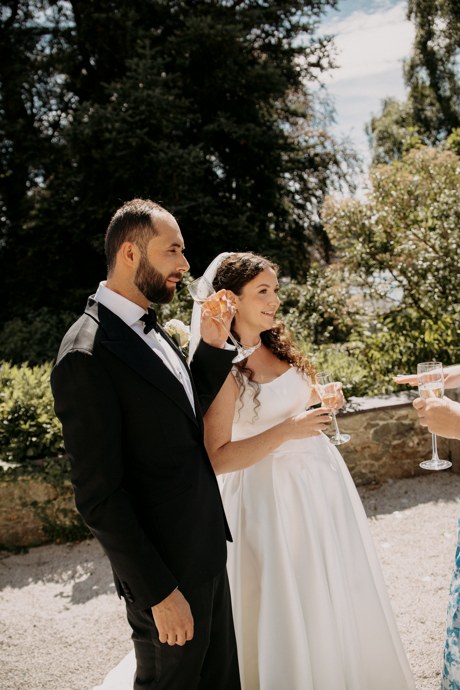 A bride and groom share a champagne toast outdoors on their wedding day, surrounded by lush greenery in warm sunlight.