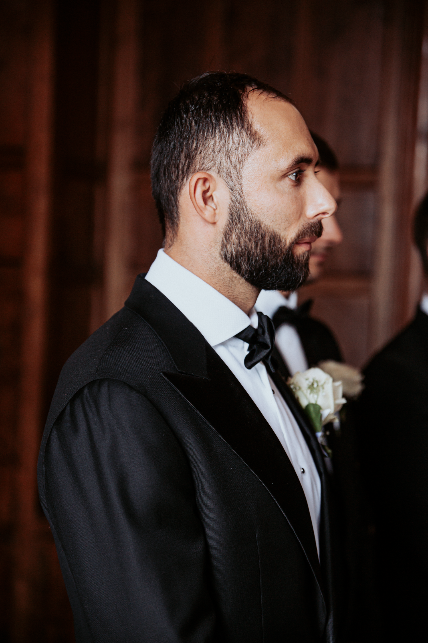 A bearded groom in a black tuxedo with bow tie and boutonniere stands in profile during a wedding ceremony.