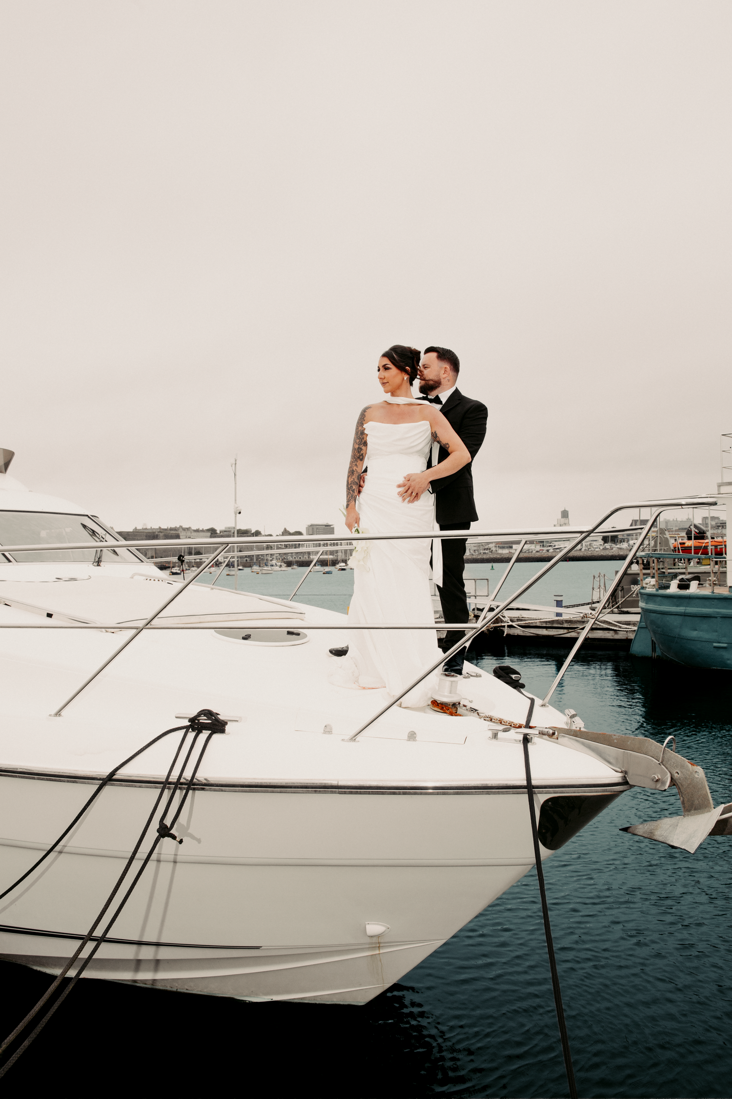 Bride and groom embracing on the bow of a white luxury yacht in a marina on their wedding day.