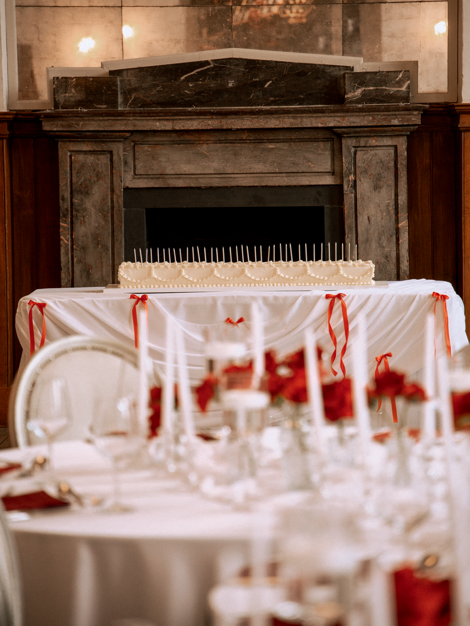 Wedding cake on draped table with red roses near ornate fireplace.