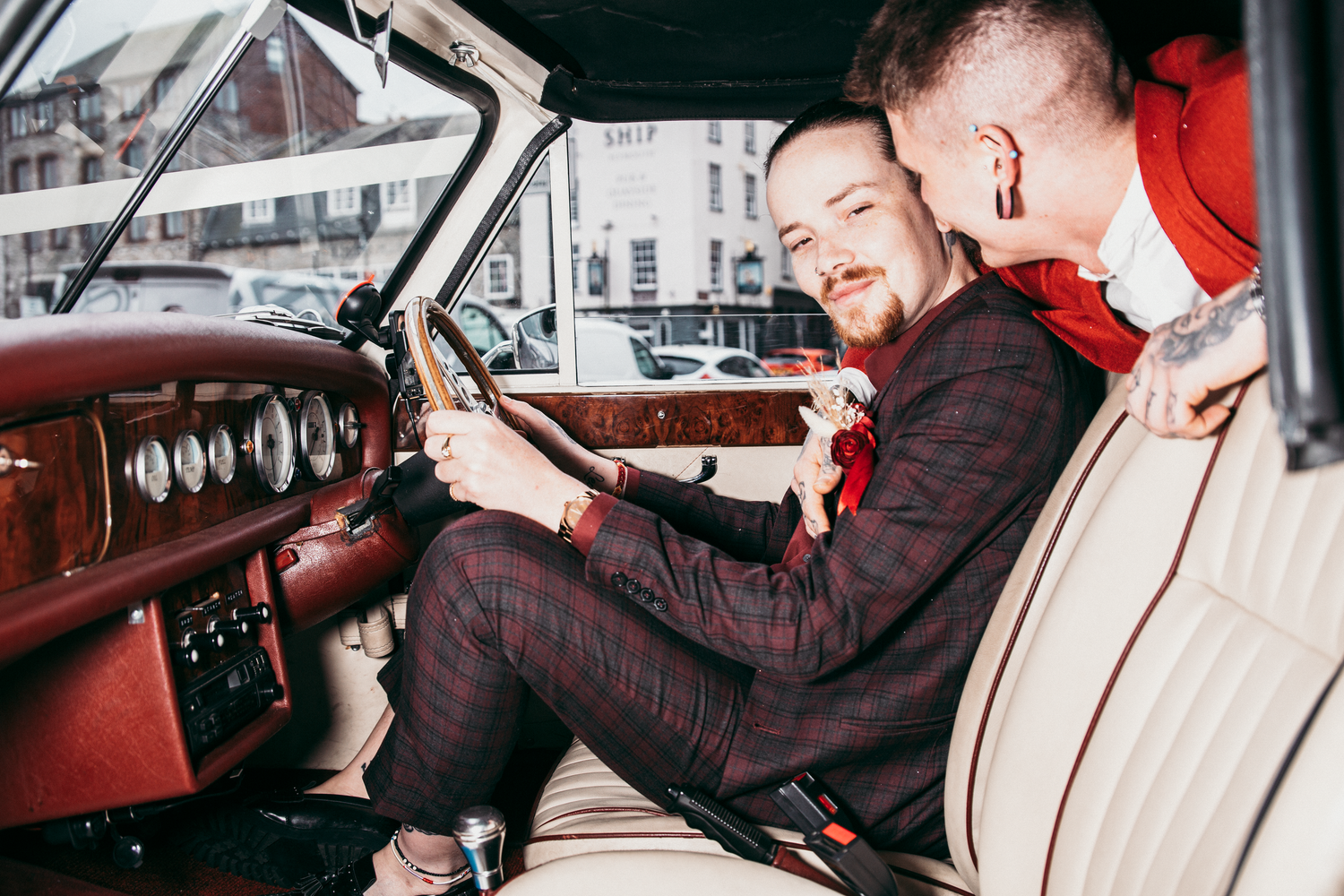 Two grooms sit inside a classic vintage car, smiling and sharing an intimate moment on their wedding day.