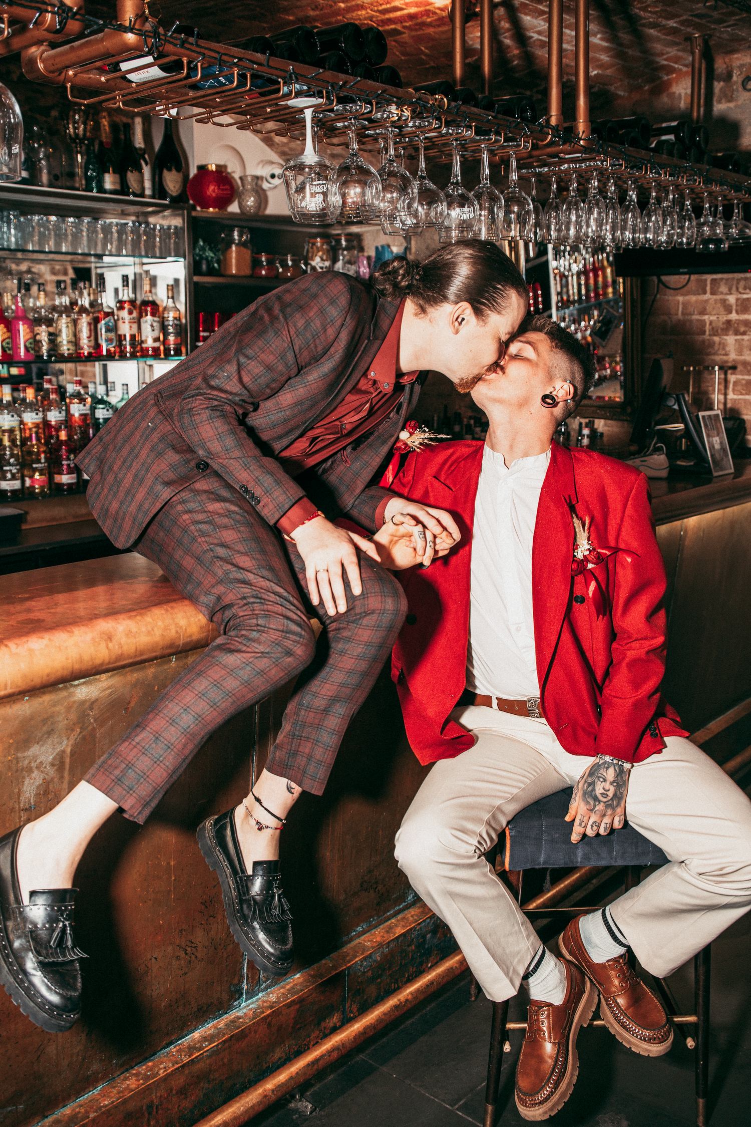 Two grooms sharing a kiss at bar counter, one in plaid suit, other in red blazer.