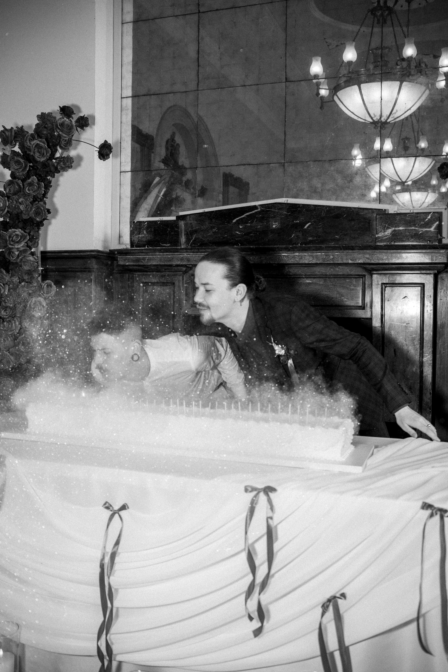 Black and white photo of groom cutting tall candle wedding cake at elegant reception venue.