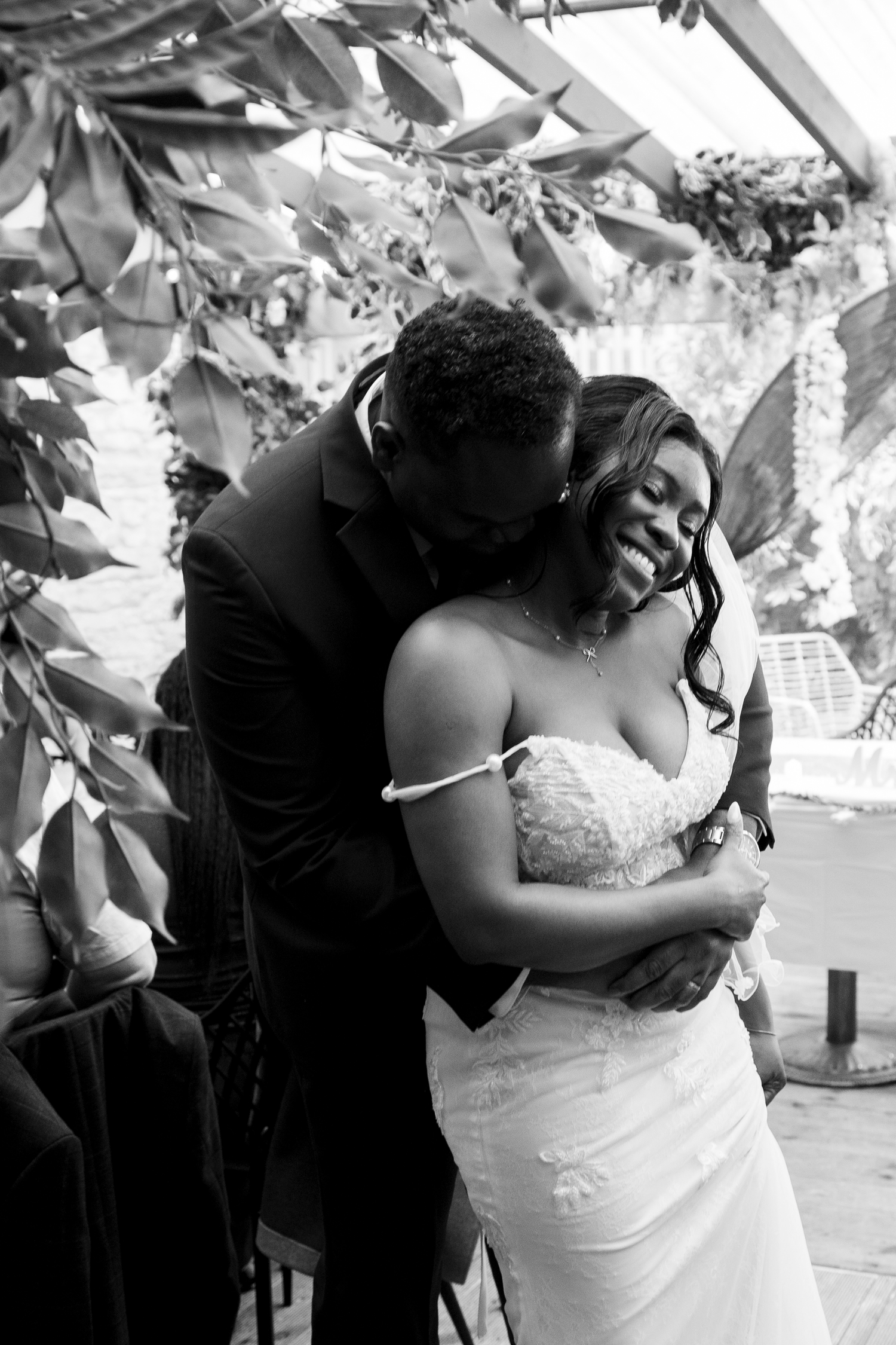 Black and white photo of a groom embracing his smiling bride during their wedding ceremony outdoors.