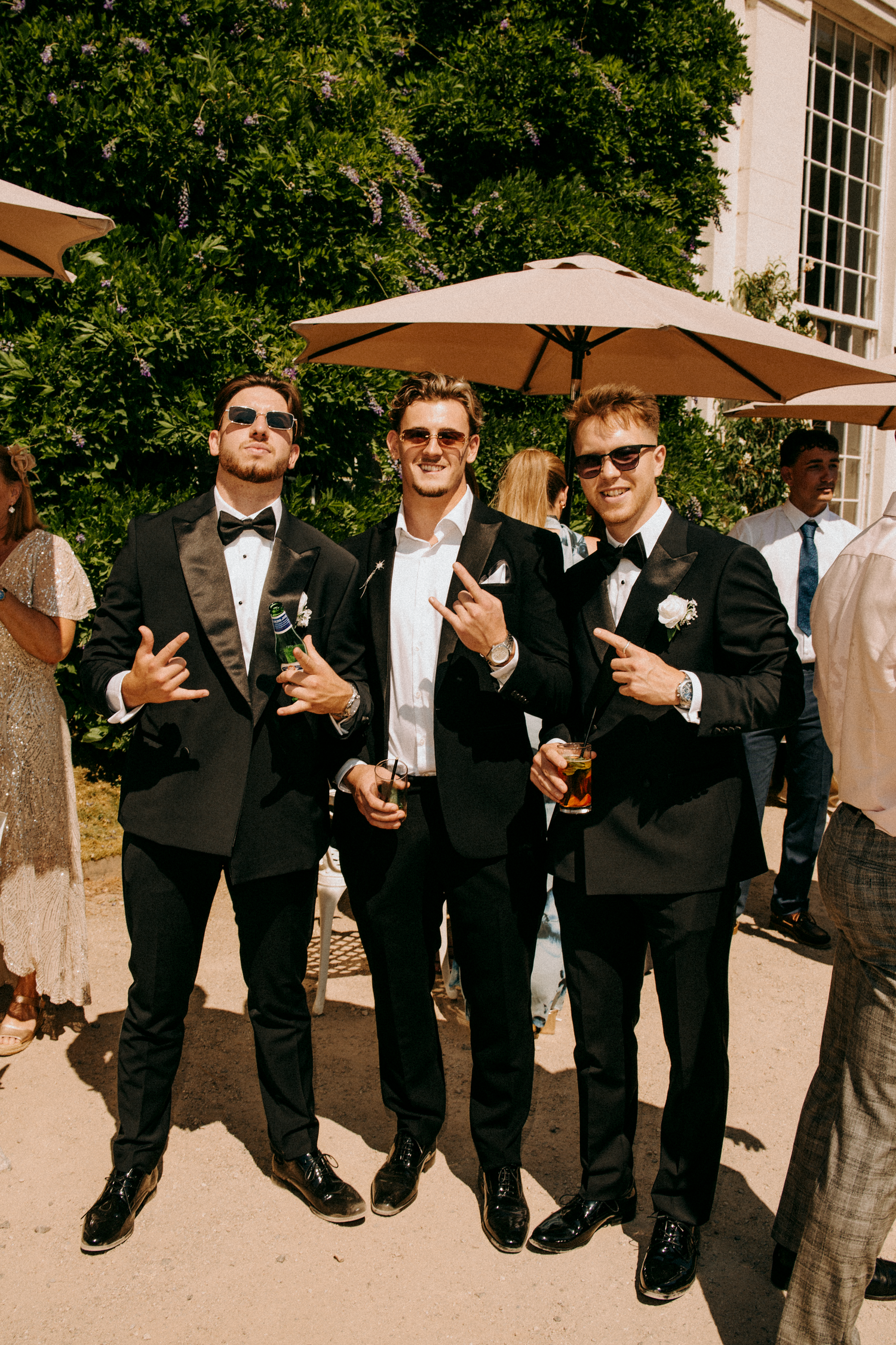 Three groomsmen in black tuxedos wearing sunglasses posing outdoors at a summer wedding celebration.