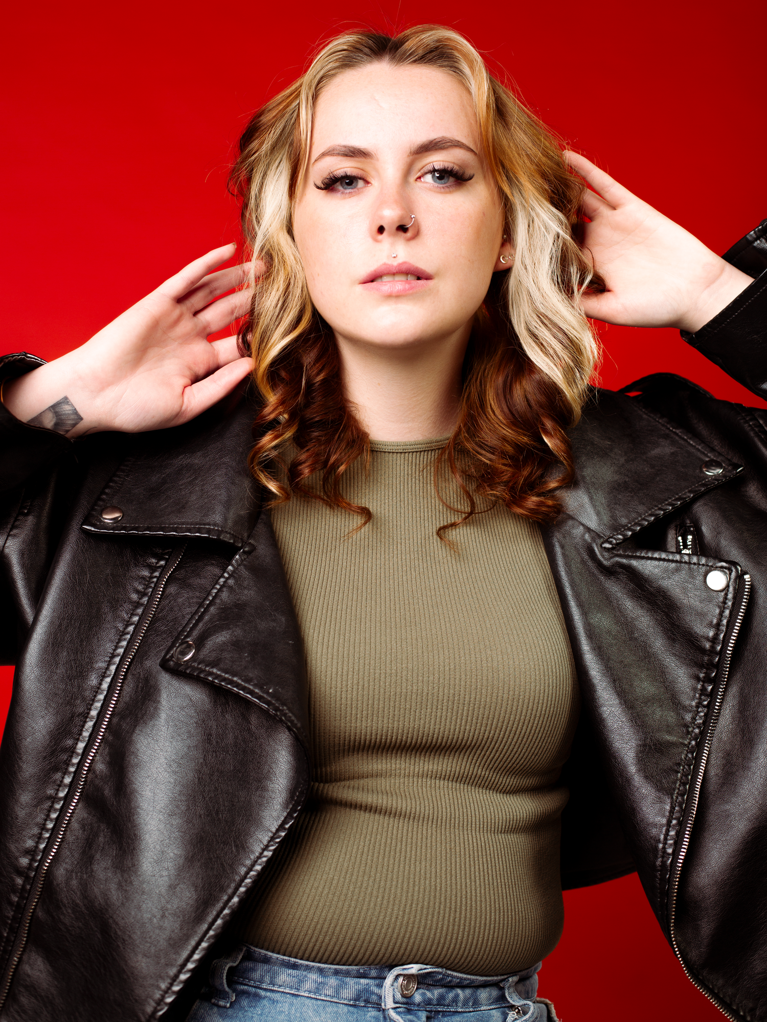 Young woman in leather jacket and olive top posing against a bold red background. Acting Headshot
