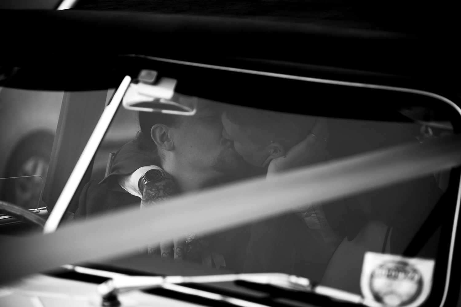 Black and white photo of two grooms sharing a kiss inside a vintage wedding car.