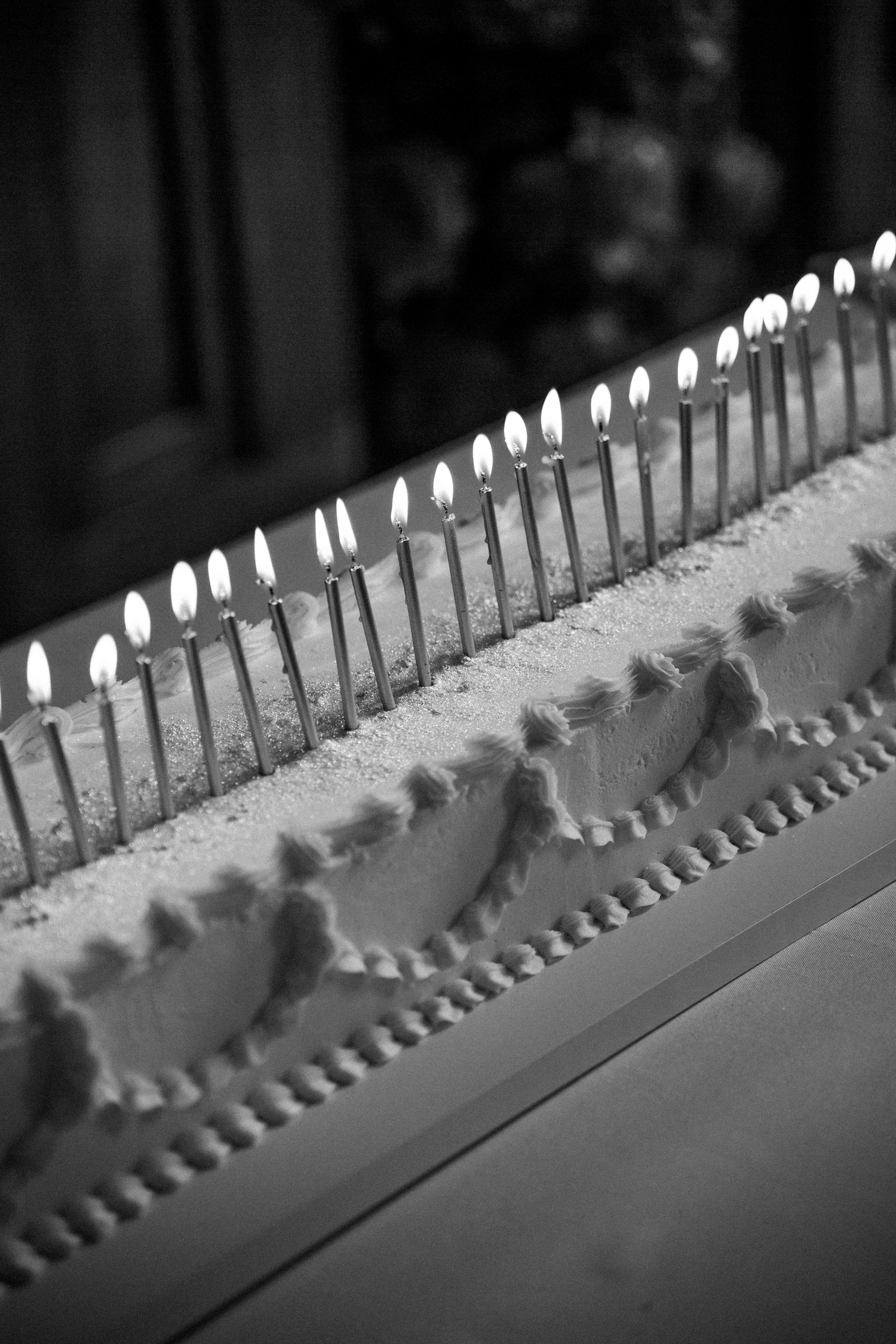 Black and white close-up of lit birthday candles on white wedding cake with decorative frosting.