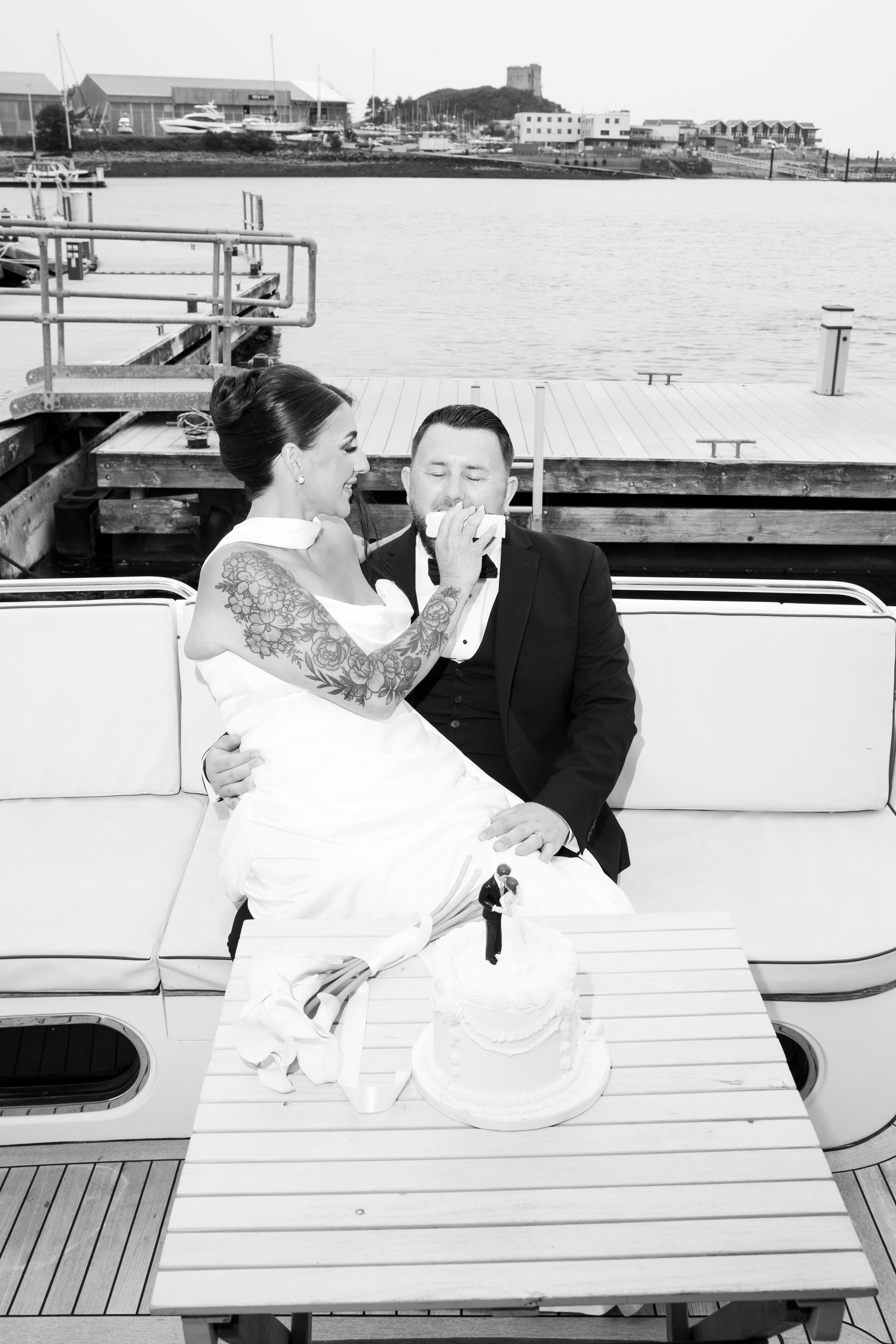 Black and white photo of newlyweds sharing cake on a yacht deck, laughing together by the water.