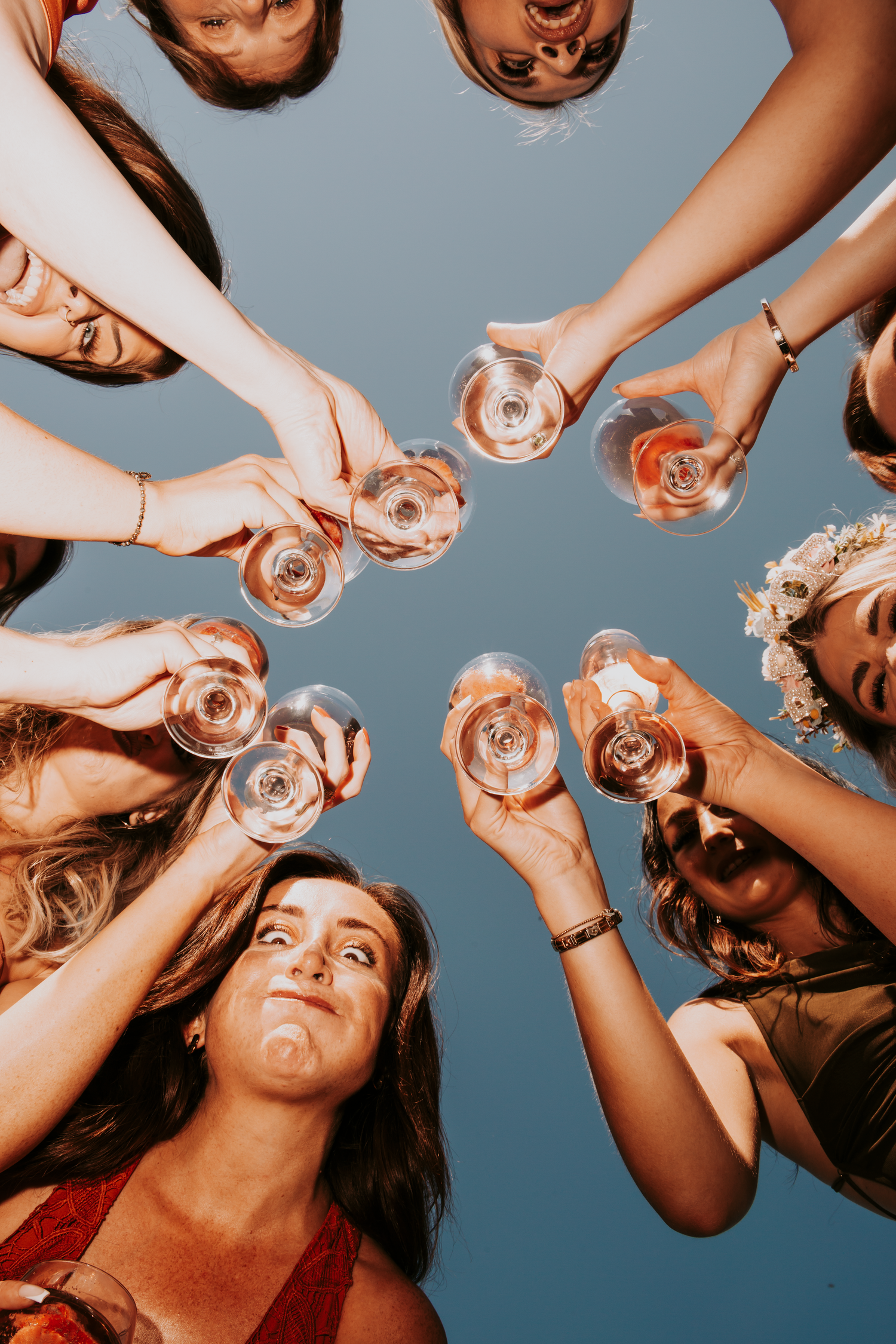 Wedding guests clink wine glasses in a circle toast photographed from below against blue sky.