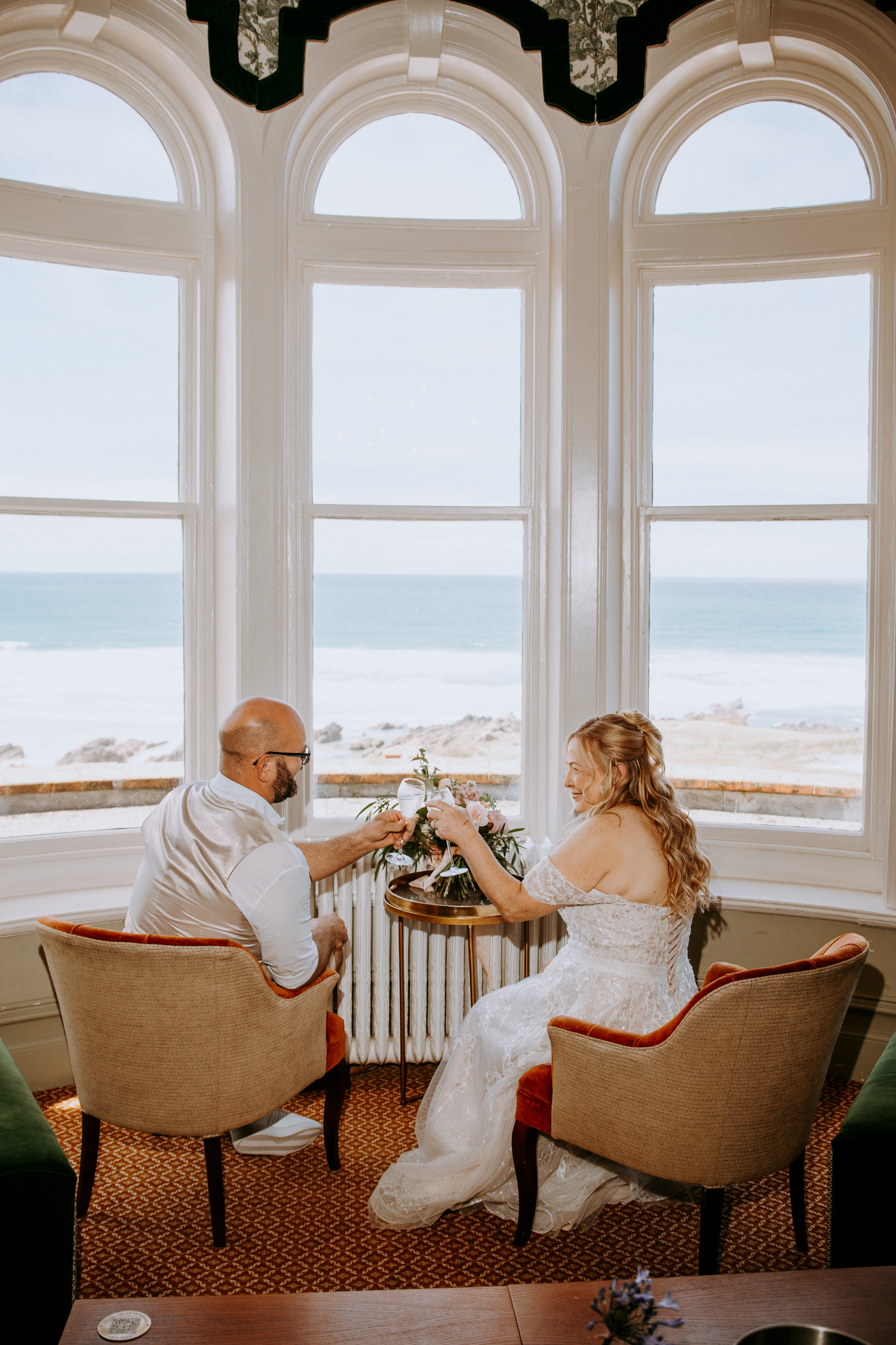 A bride and groom toast with flowers by large arched windows overlooking a scenic ocean view.
