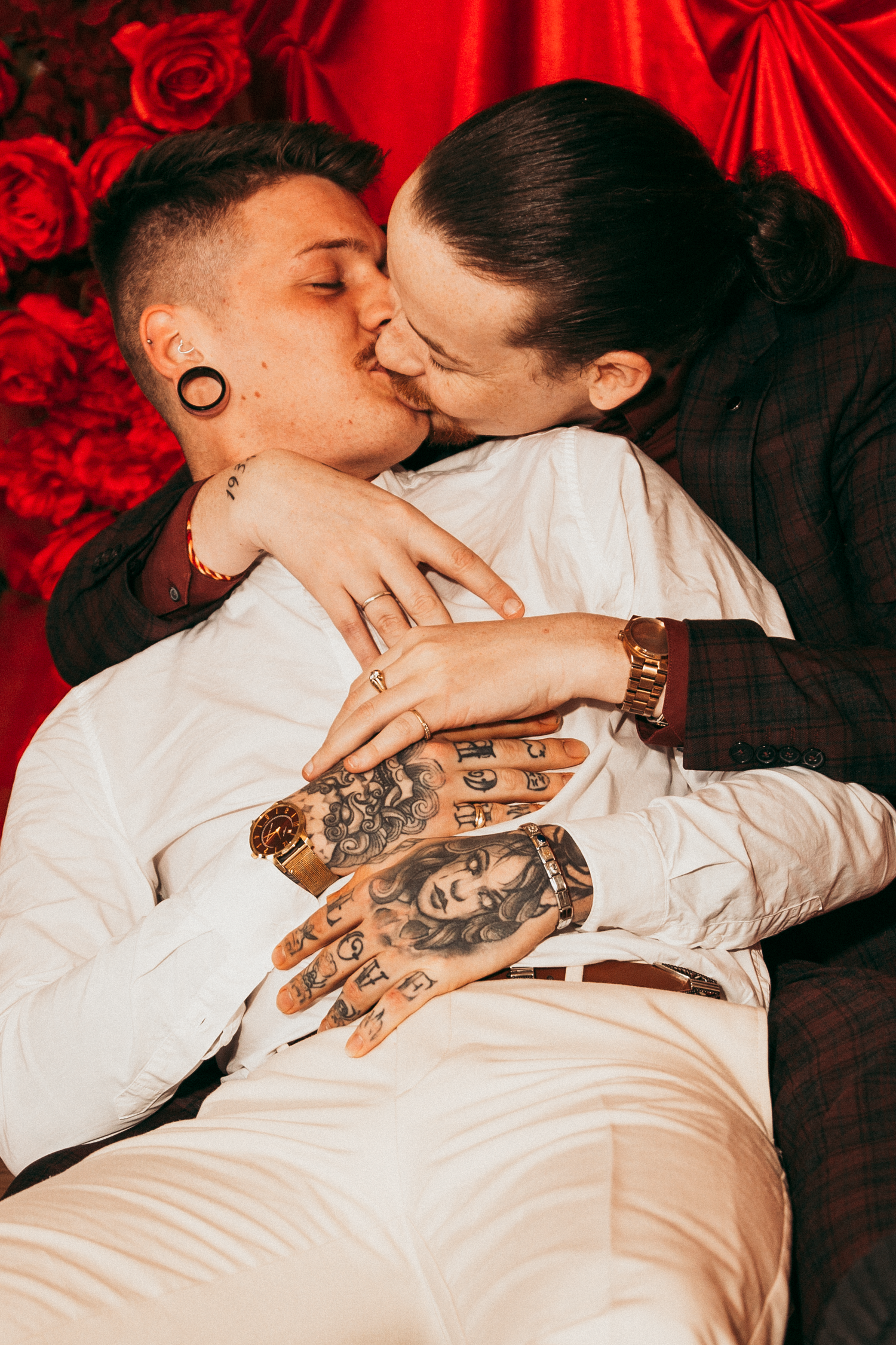 Couple sharing romantic kiss in photo booth with red rose backdrop, showing tattooed hands.