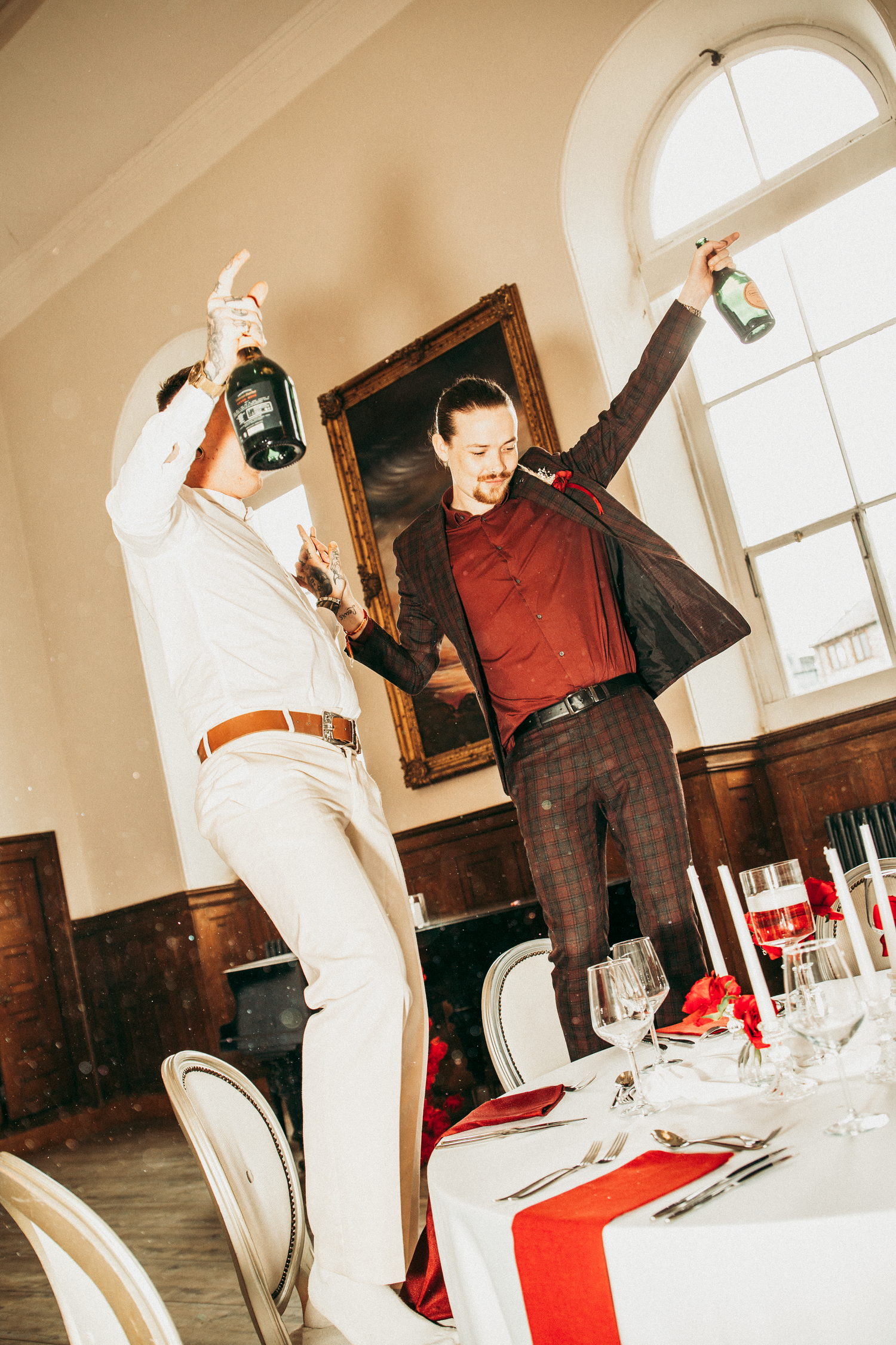 Two grooms celebrate joyfully, holding champagne bottles aloft in an elegant wedding reception hall.