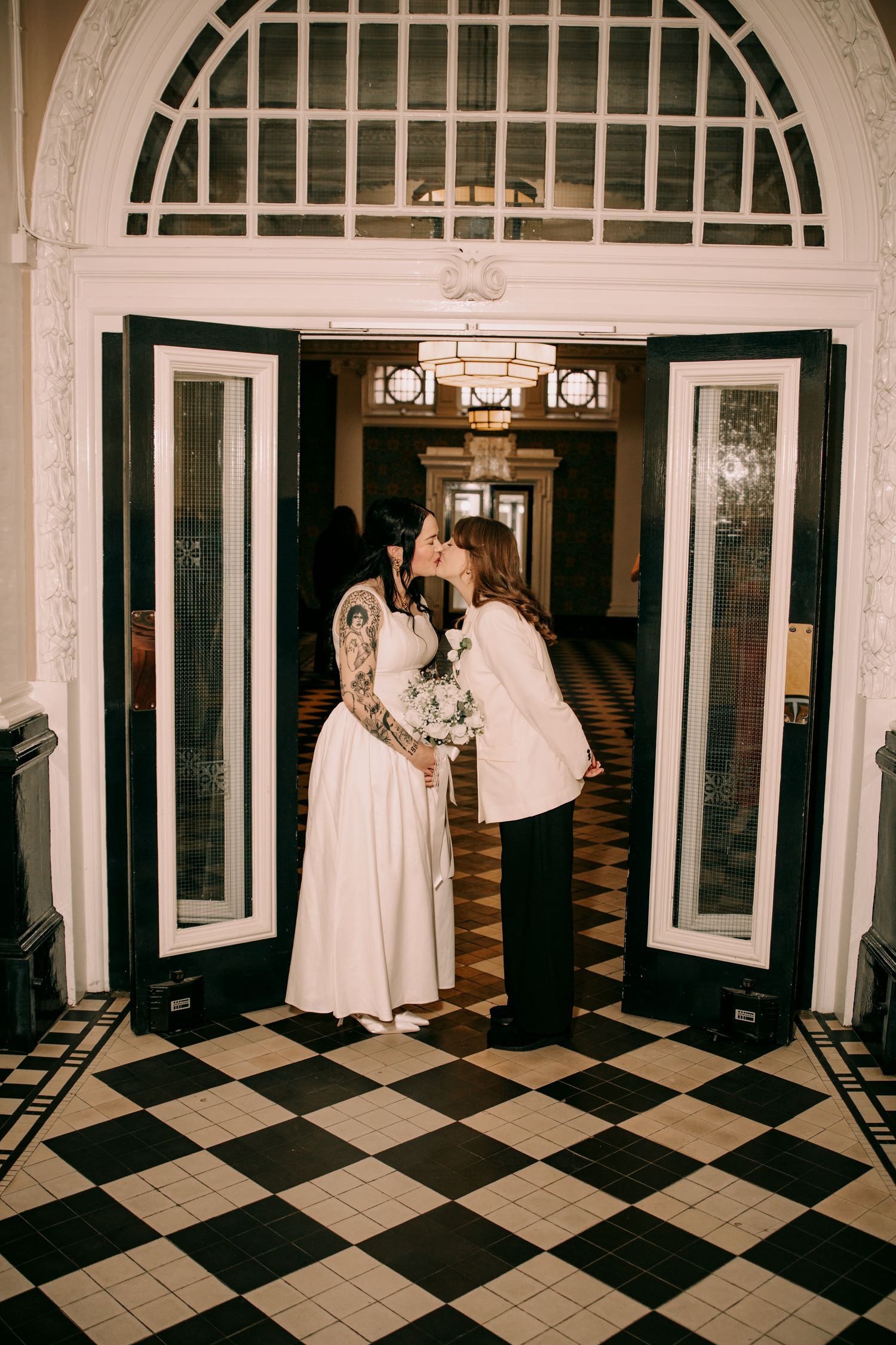 Two brides sharing a kiss in elegant black and white checkered foyer doorway with arched window above.