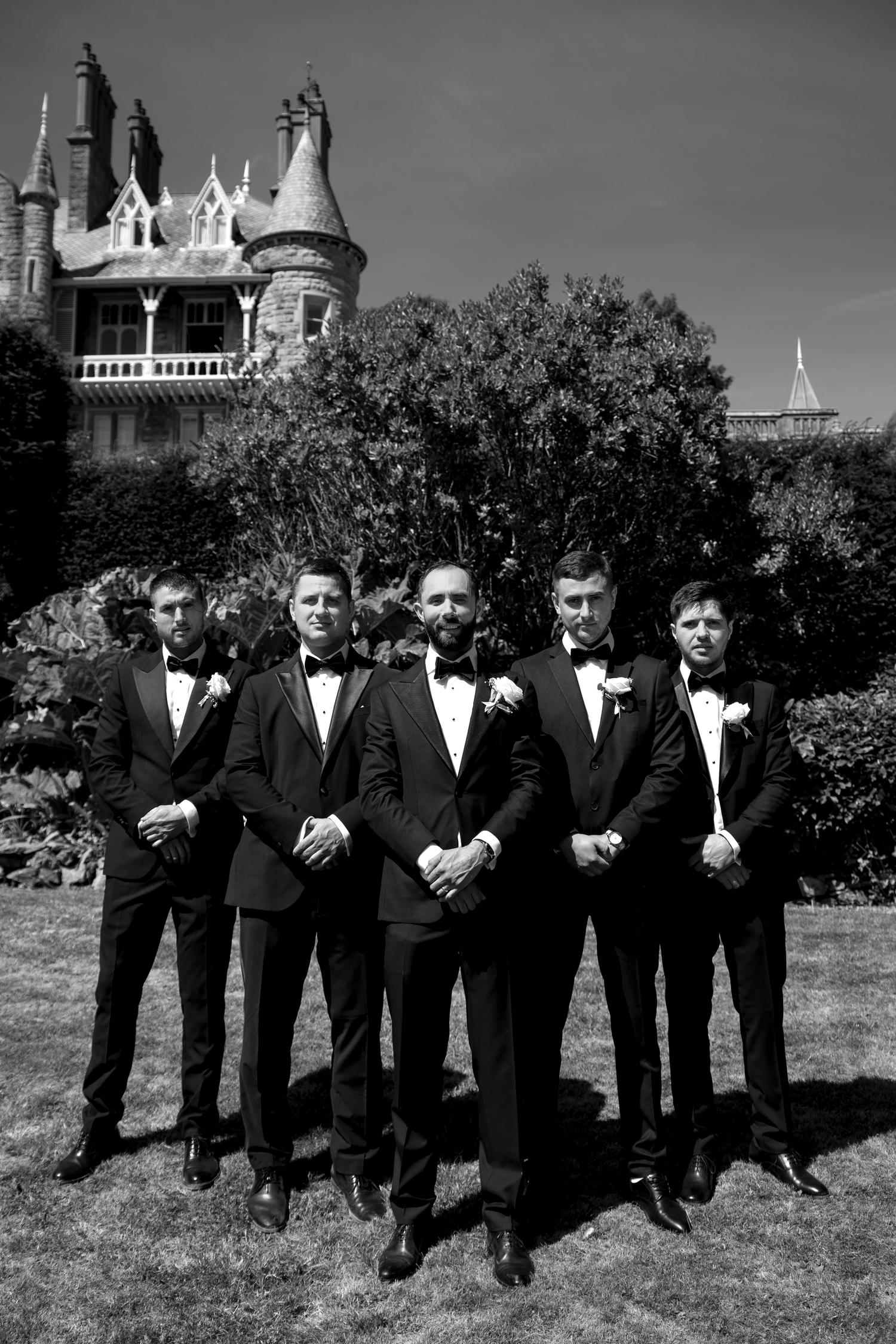 Five groomsmen in black tuxedos with bow ties pose formally outdoors before a Gothic-style mansion in black and white photo.