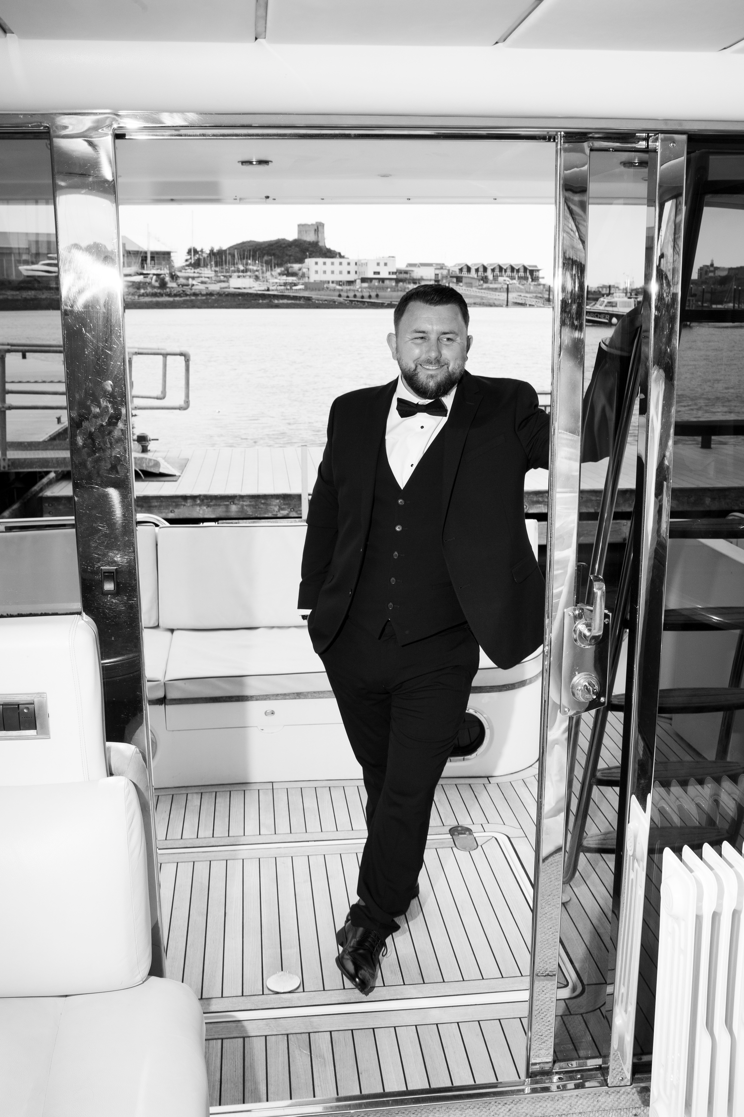 Dapper groom in black tuxedo with bow tie posing in yacht doorway with marina views, black and white.