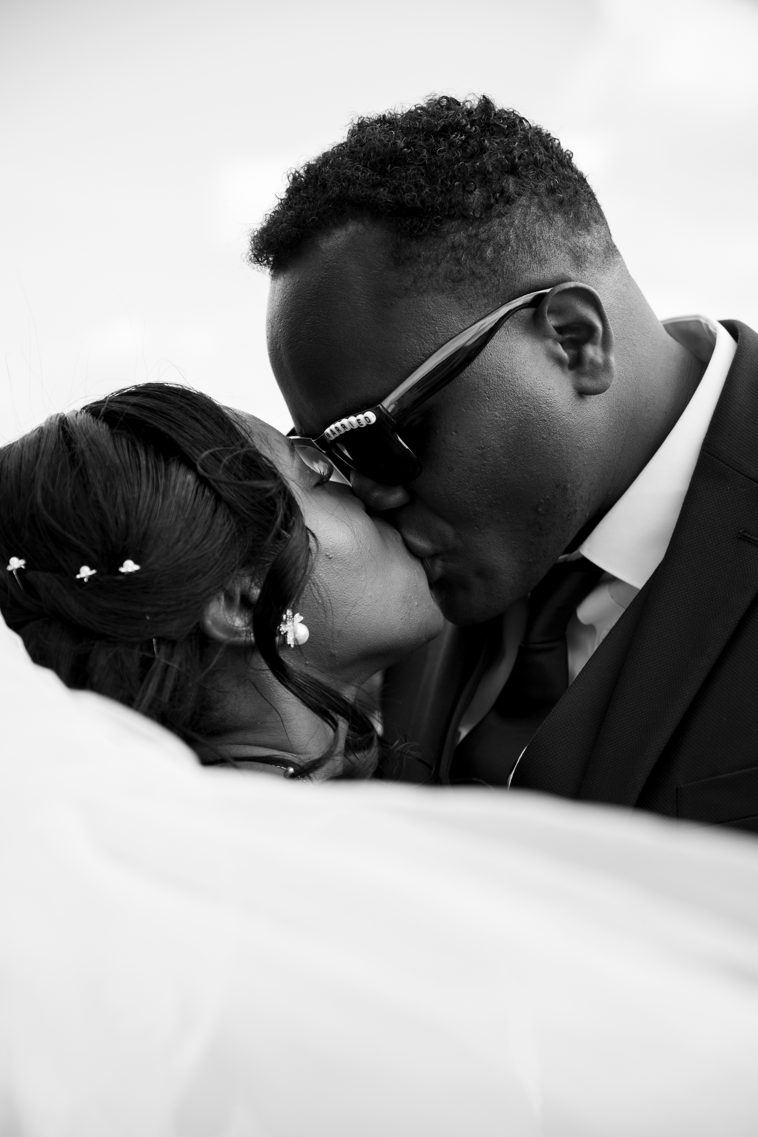 A newlywed couple shares a tender kiss, the groom wearing sunglasses, captured in black and white.