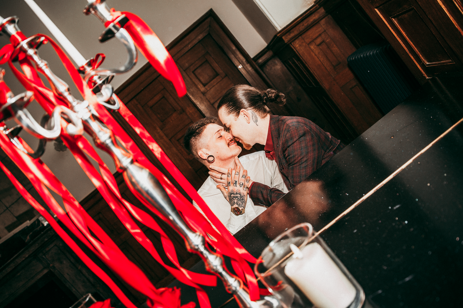 Two grooms share a first kiss at the altar surrounded by red ribbon chandelier decorations and candles.