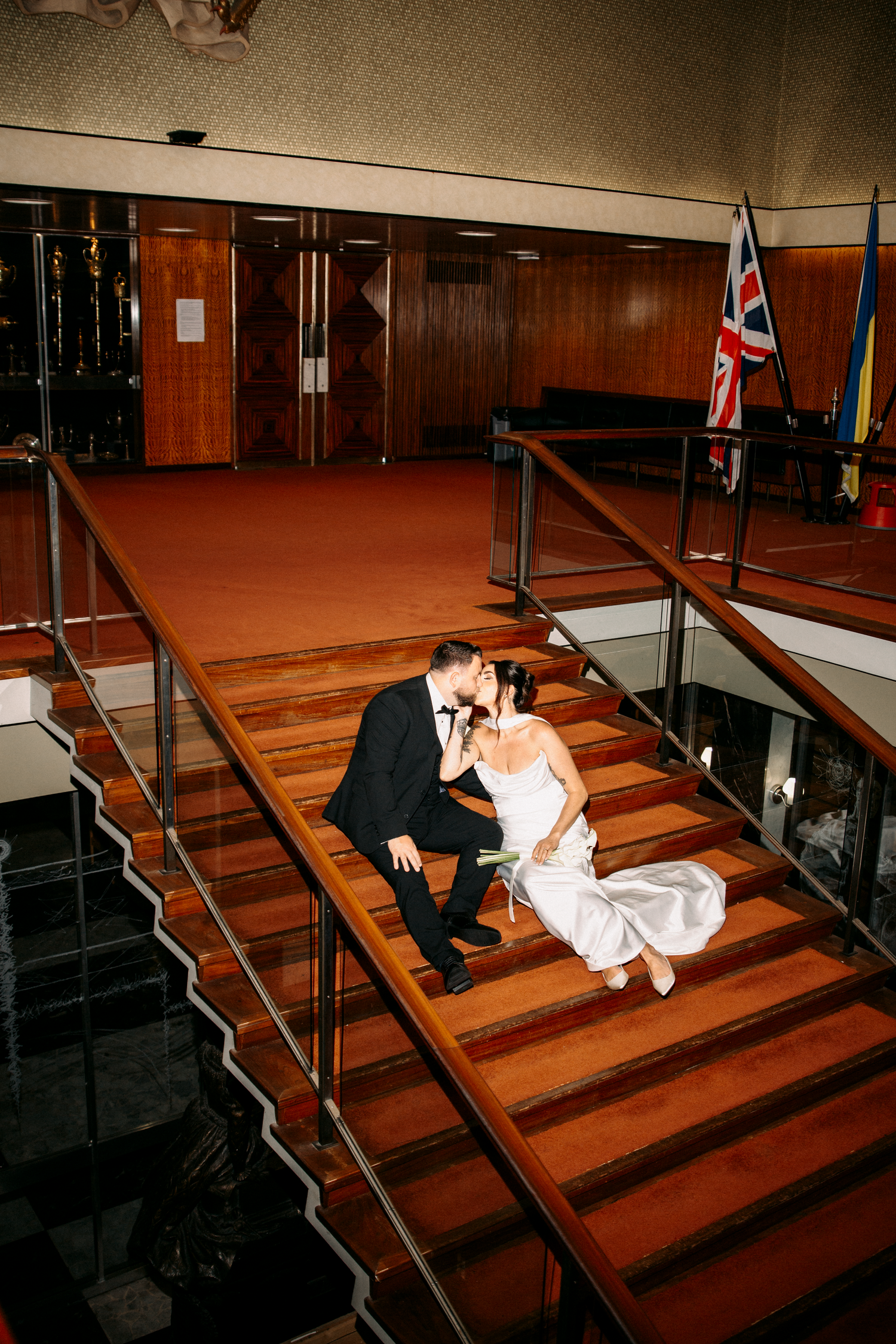 Bride and groom sharing a kiss while seated on grand interior staircase with Union Jack flag in background.