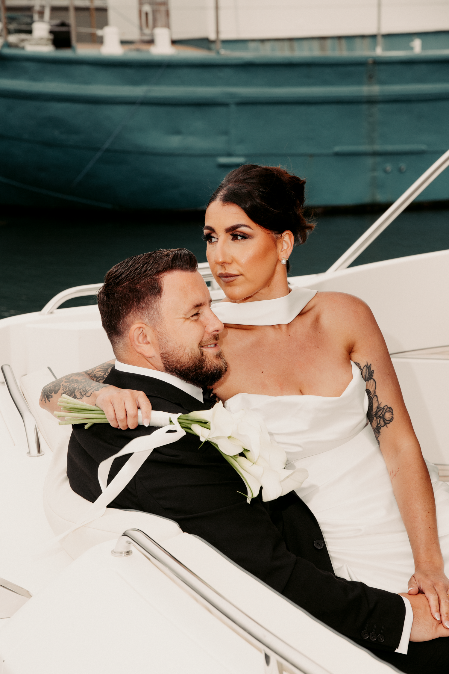 Bride in white halter gown and groom in tuxedo relaxing together on a luxury yacht at marina.
