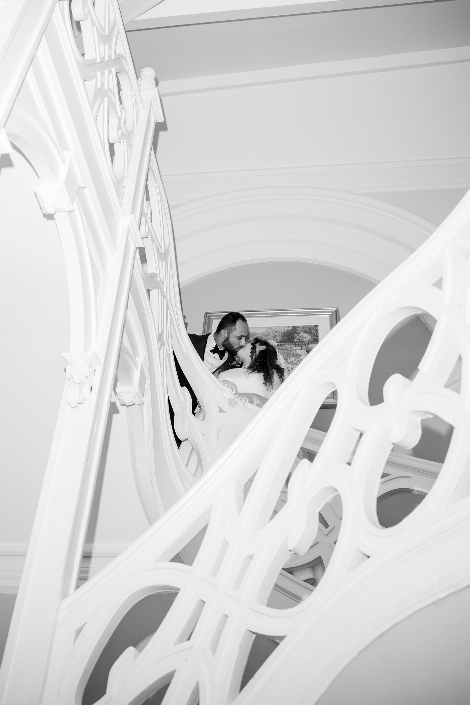 Black and white photo of a wedding couple embracing on an elegant staircase with ornate white railings.