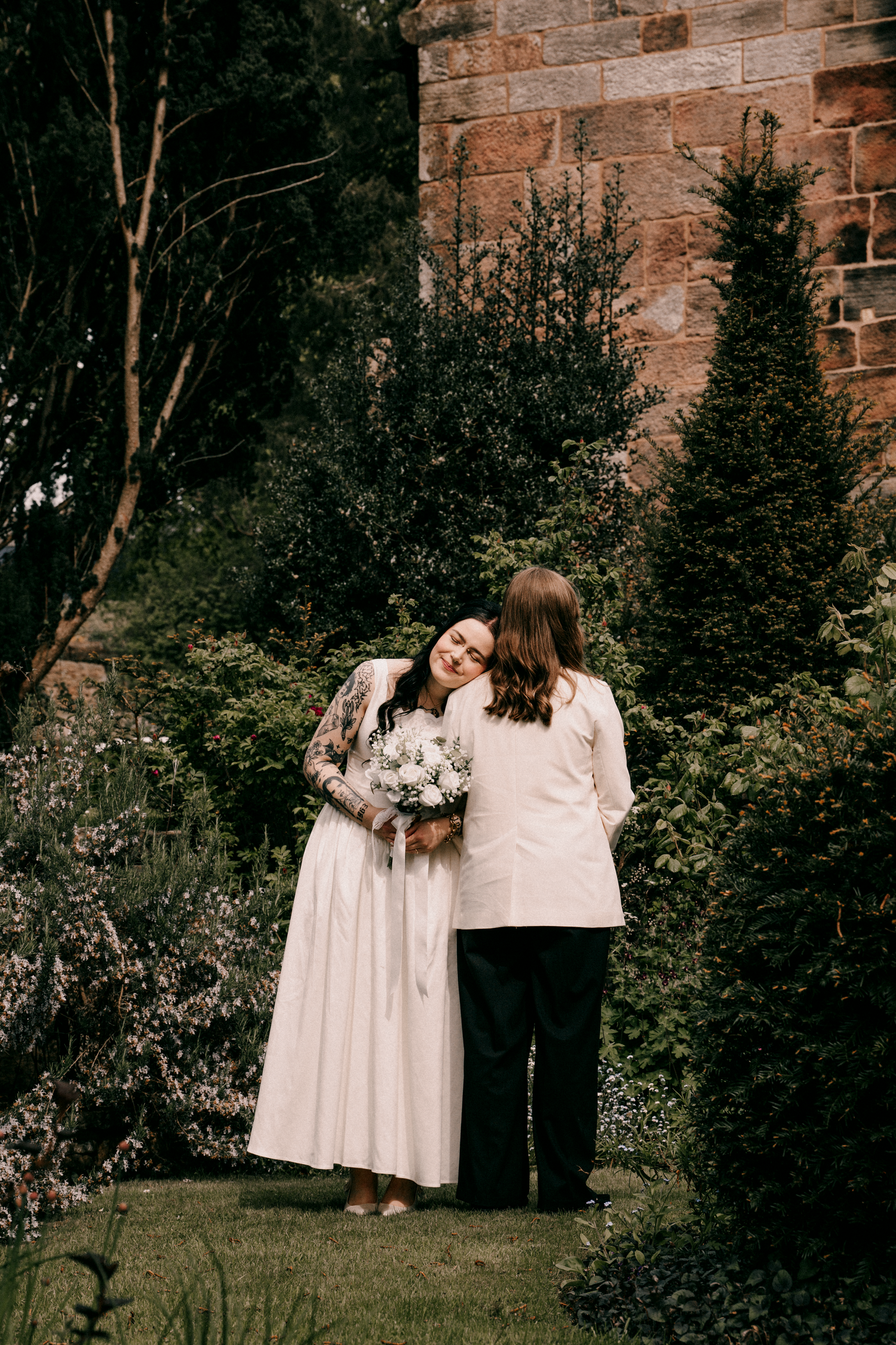 Two brides embracing in a lush garden beside a stone building, one in white dress with tattoos.