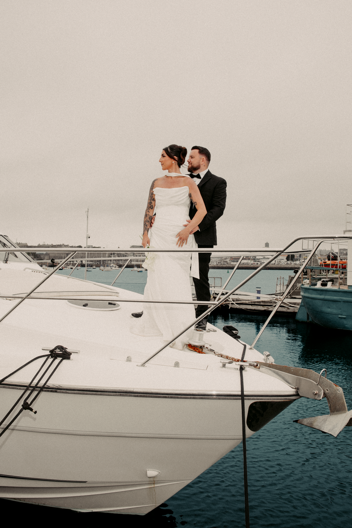 Bride and groom stand romantically on luxury yacht deck in harbor on overcast wedding day.