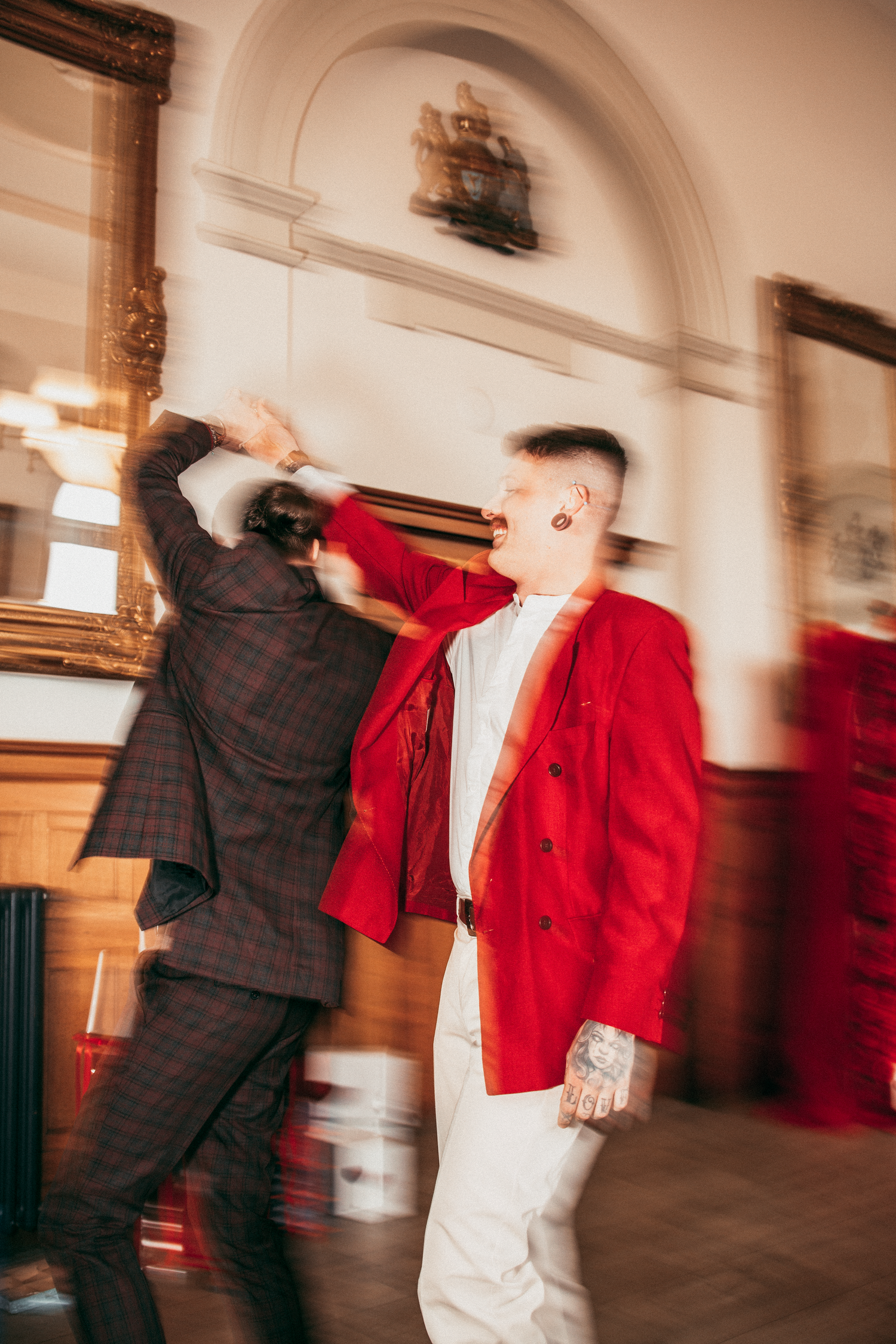 Two men dancing energetically in motion-blurred photo, one in red jacket and white pants at wedding.