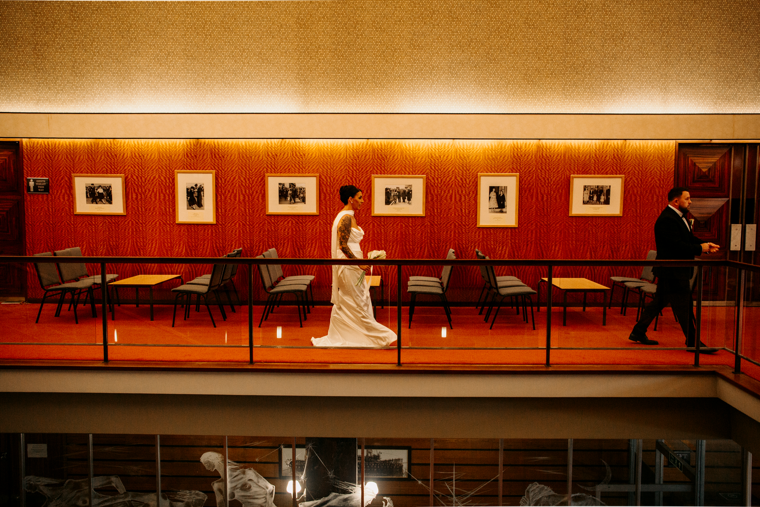 Bride and groom walk separately along a mezzanine level corridor in a sophisticated wedding venue.