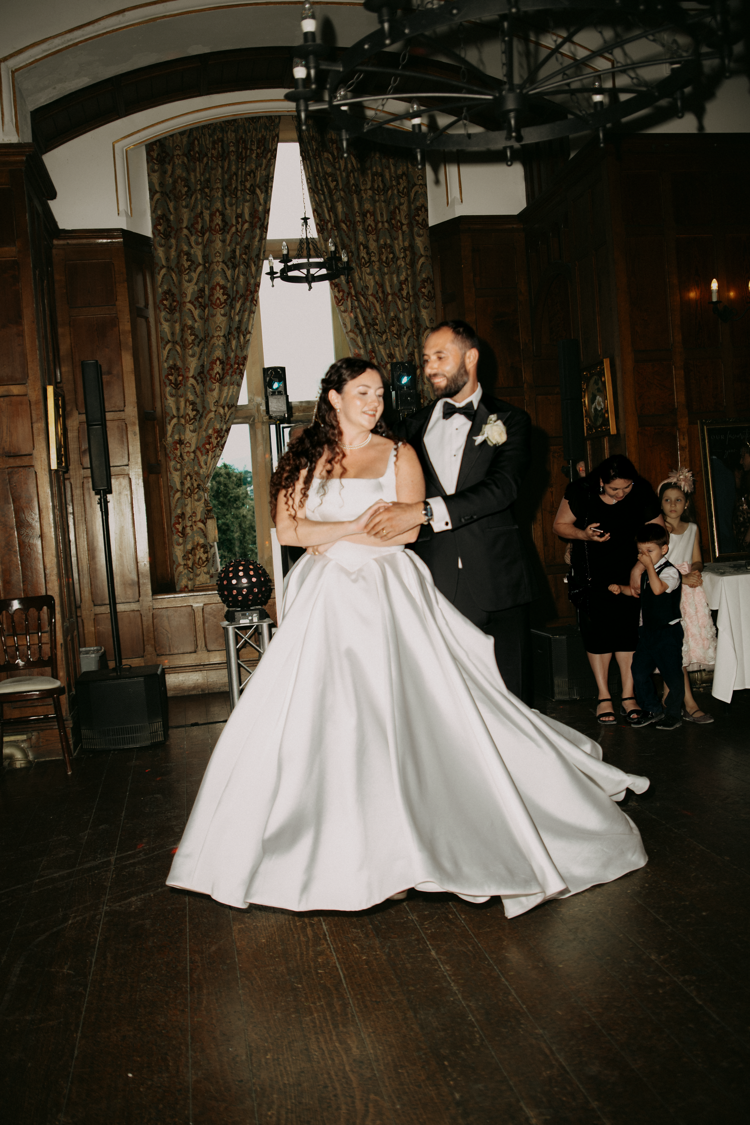 Bride and groom share their first dance in an elegant ballroom, she wears a flowing white satin gown.