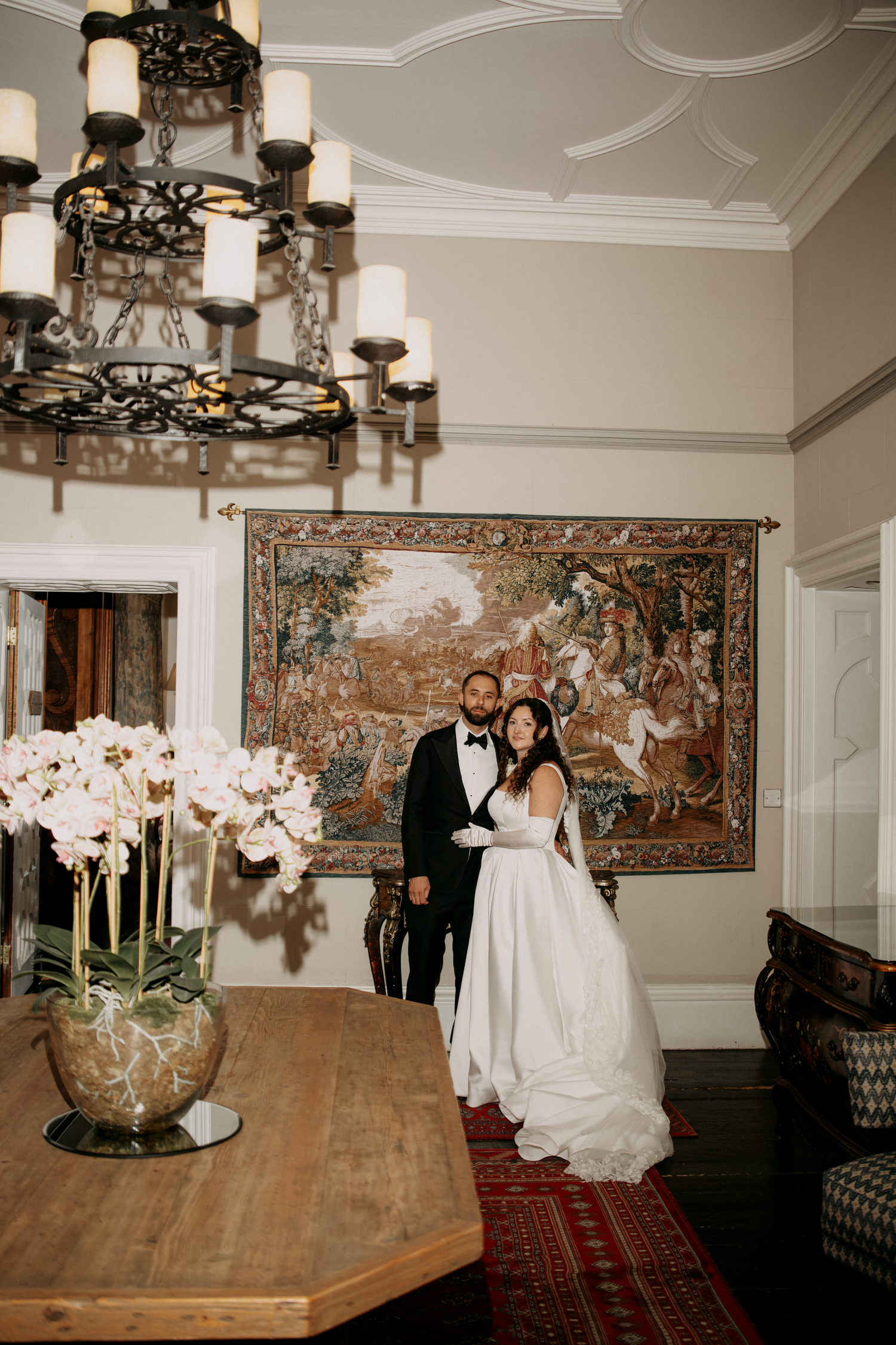 Elegant bride and groom posing in a grand room with a tapestry, chandelier, and floral arrangement on a wooden table.