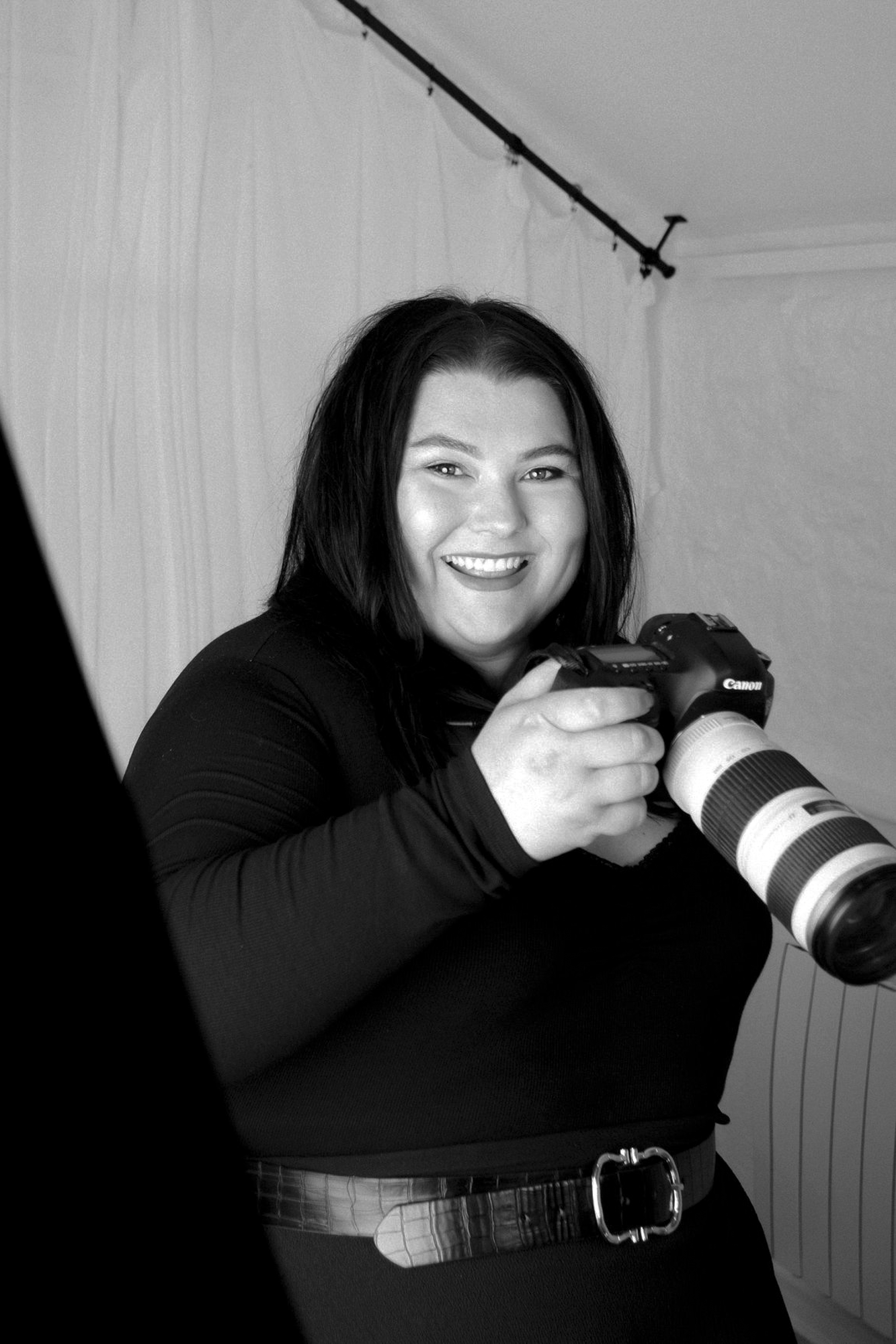 Professional female photographer smiling while holding a Canon DSLR camera with telephoto lens in black and white.